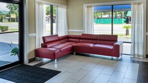 A red sectional sofa in a hotel lobby with a black doormat, beige walls, and large windows with white curtains. - Rodeway Inn Panama City (Panama City)