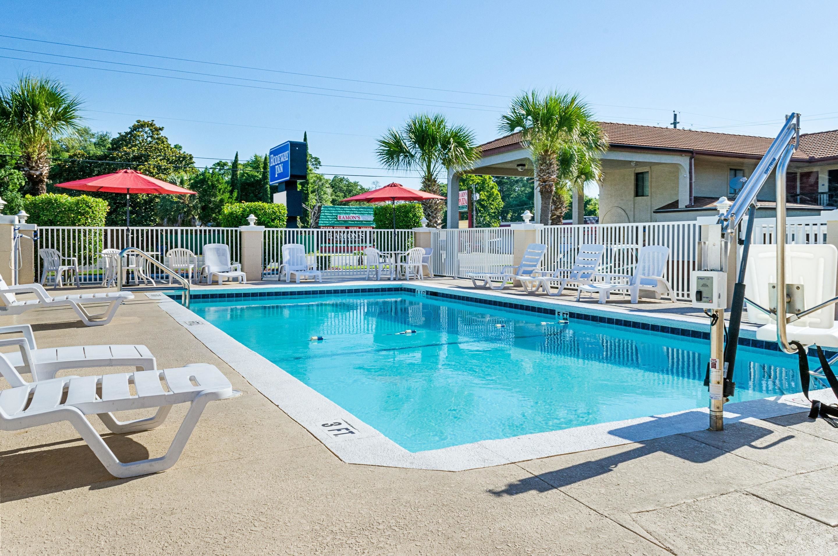 A swimming pool with lounge chairs and umbrellas, surrounded by a fence and palm trees.
