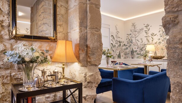 A room with a stone wall, a mirror, a table with a lamp, a vase with flowers, and blue chairs.