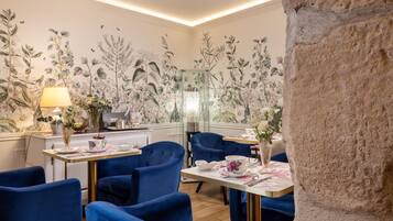 A dining area with blue upholstered chairs, a table with floral tablecloth, and a wall with botanical wallpaper.