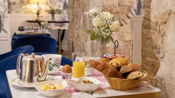 Een ontbijttafel met een theepot, kopjes, een glas sinaasappelsap, een fruitschaal, een broodmand en een vaas met bloemen.