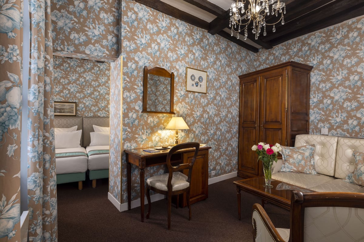 A room with floral wallpaper, a wooden cabinet, a sofa, a desk with a chair, and a chandelier.