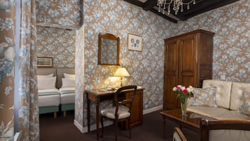 Ein Zimmer mit blumigem Wandpapier, einem Holzschrank, einem Sofa, einem Schreibtisch mit Stuhl und einer Kandelaber.