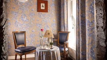 A room with two chairs, a table with a glass top, a vase with a flower, a bottle, and a framed picture on the wall.