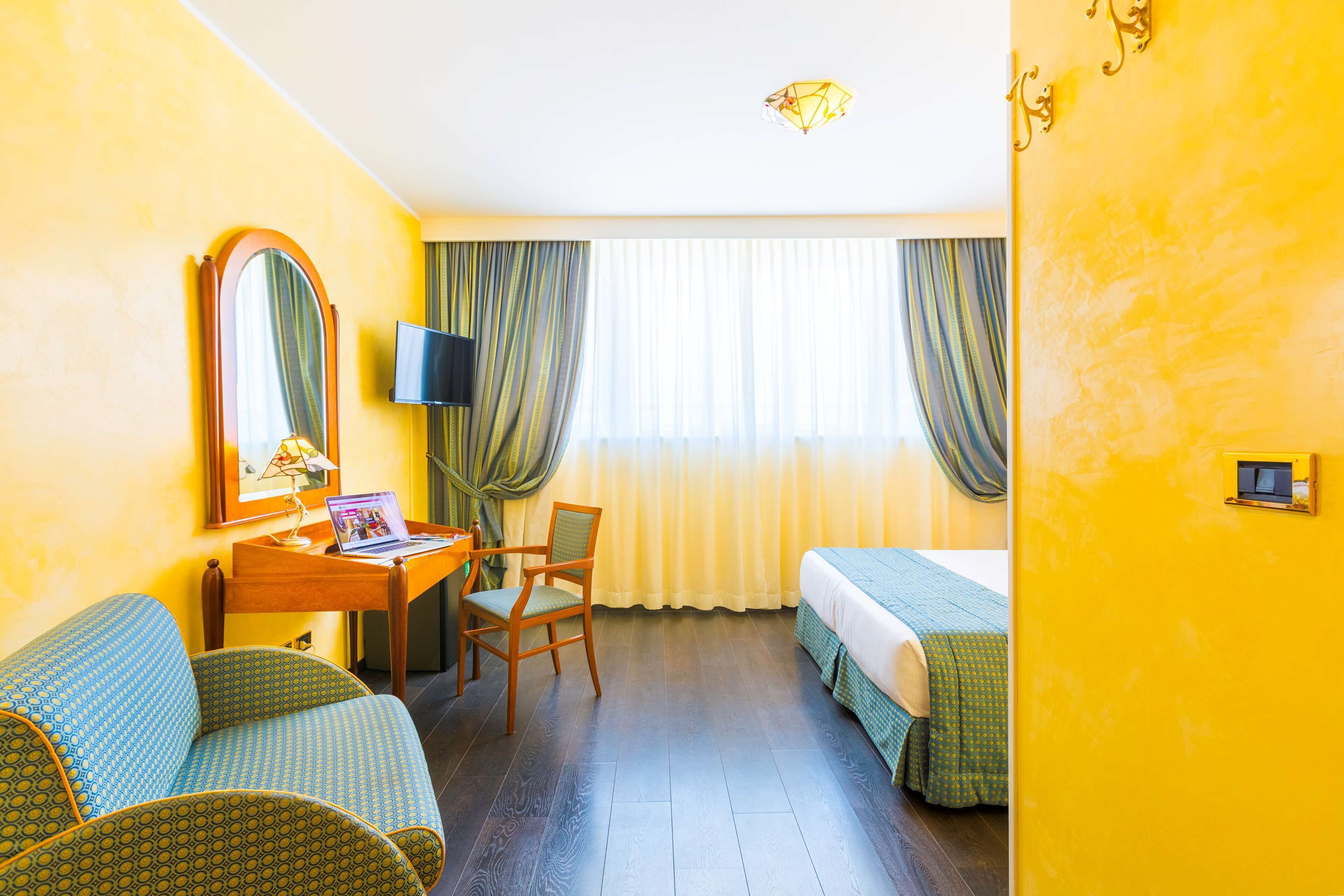 Una habitación de hotel con una cama, un escritorio con una computadora portátil, una silla, un televisor y un espejo grande.