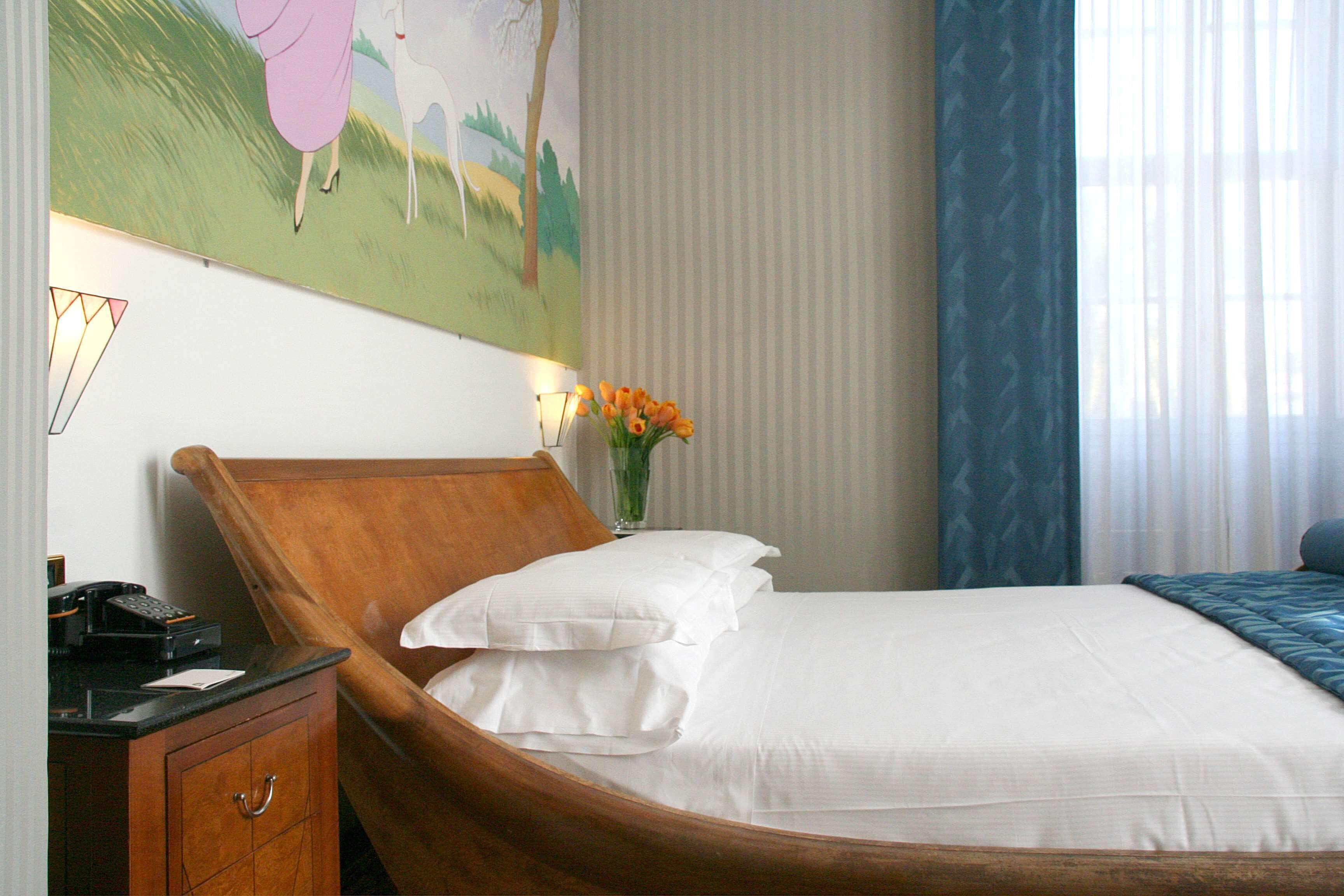 A wooden bed with white linens, a bedside table with a lamp, and a painting of a landscape on the wall.