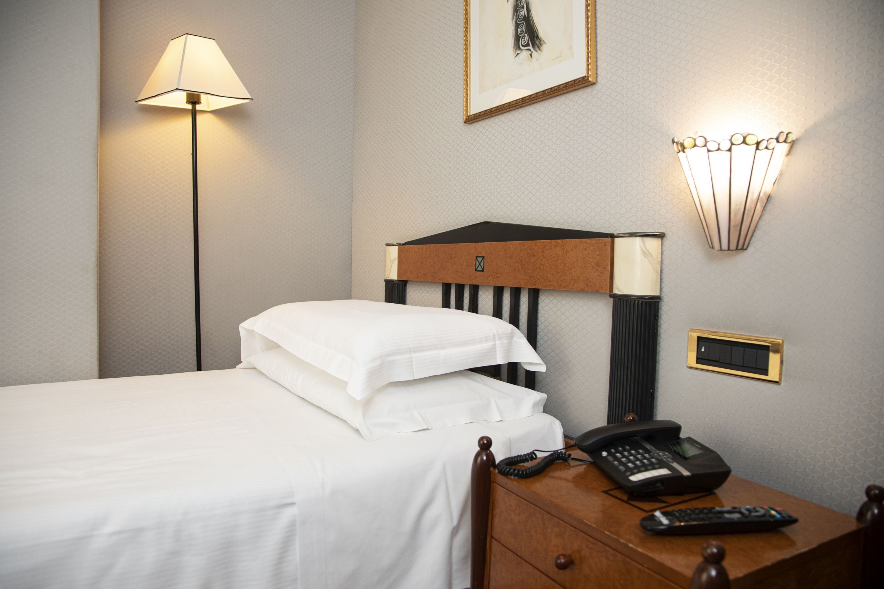 A hotel room with a bed, bedside lamp, bedside table with a phone and remote control, and a wall-mounted light fixture.
