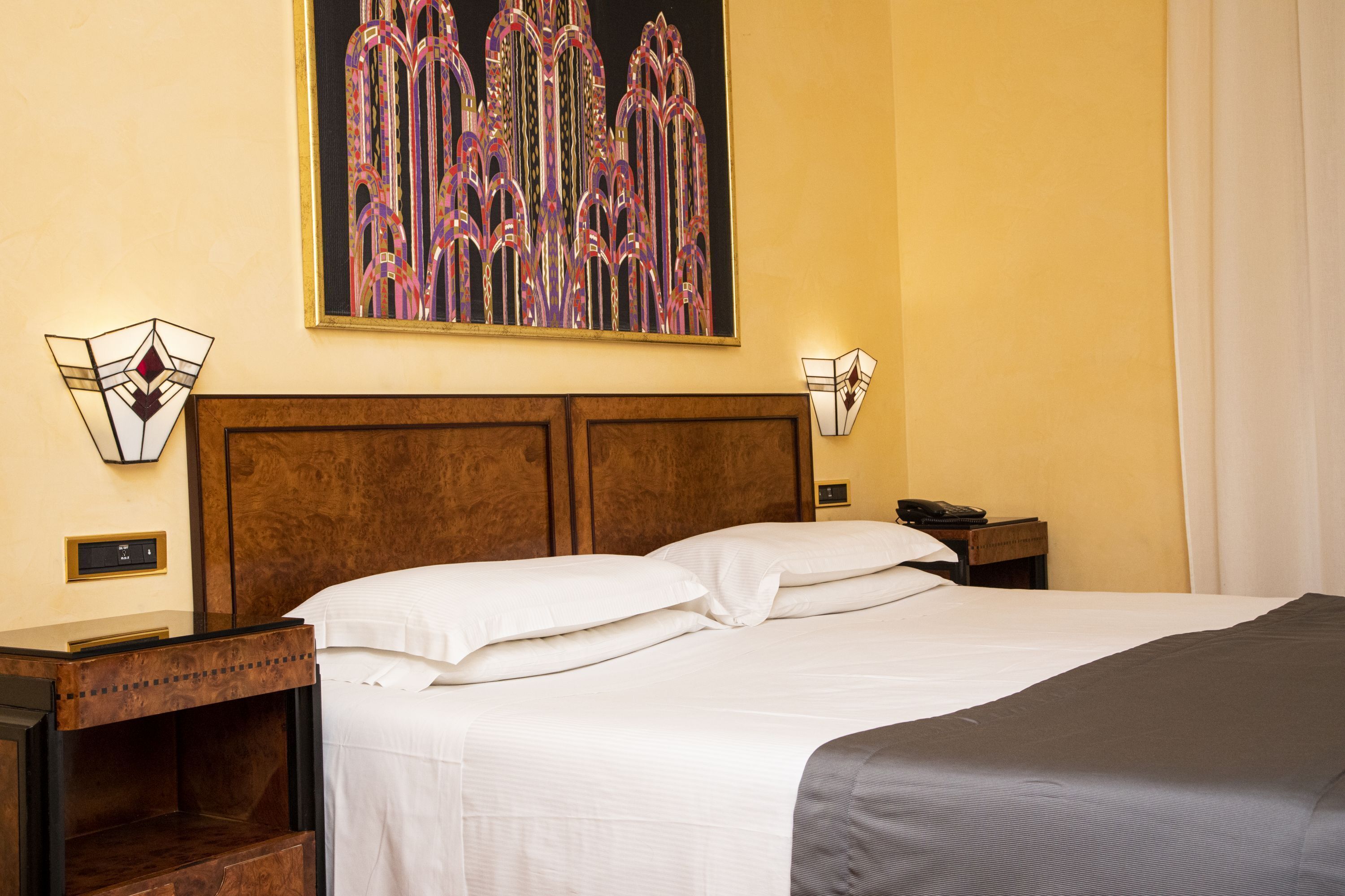 A hotel room with a large bed, wooden headboard, two bedside tables, a nightstand, a wall-mounted lamp, and a framed artwork above the bed.