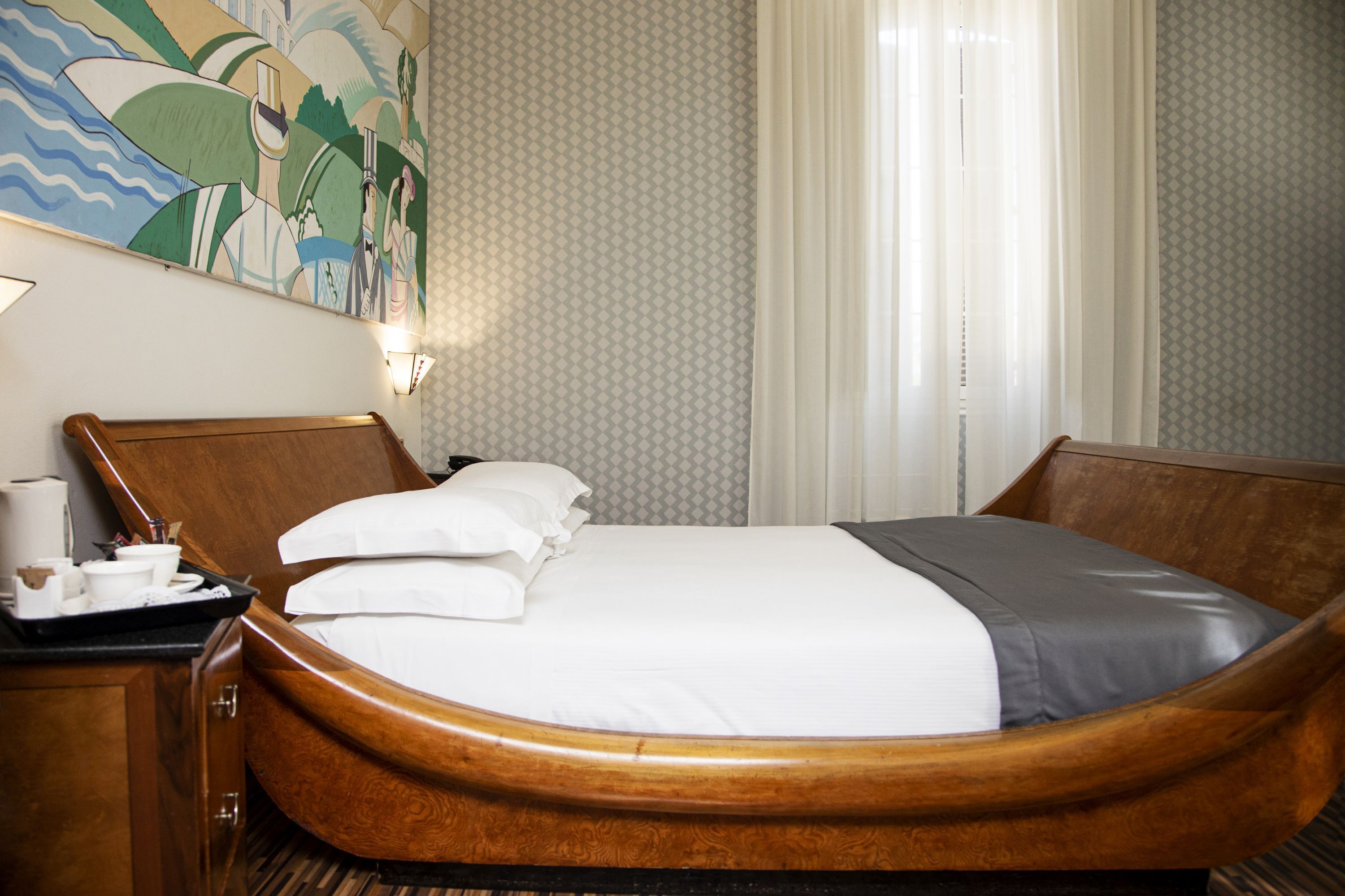 A hotel room with a wooden bed, a nightstand with a tray of toiletries, and a large abstract painting on the wall.