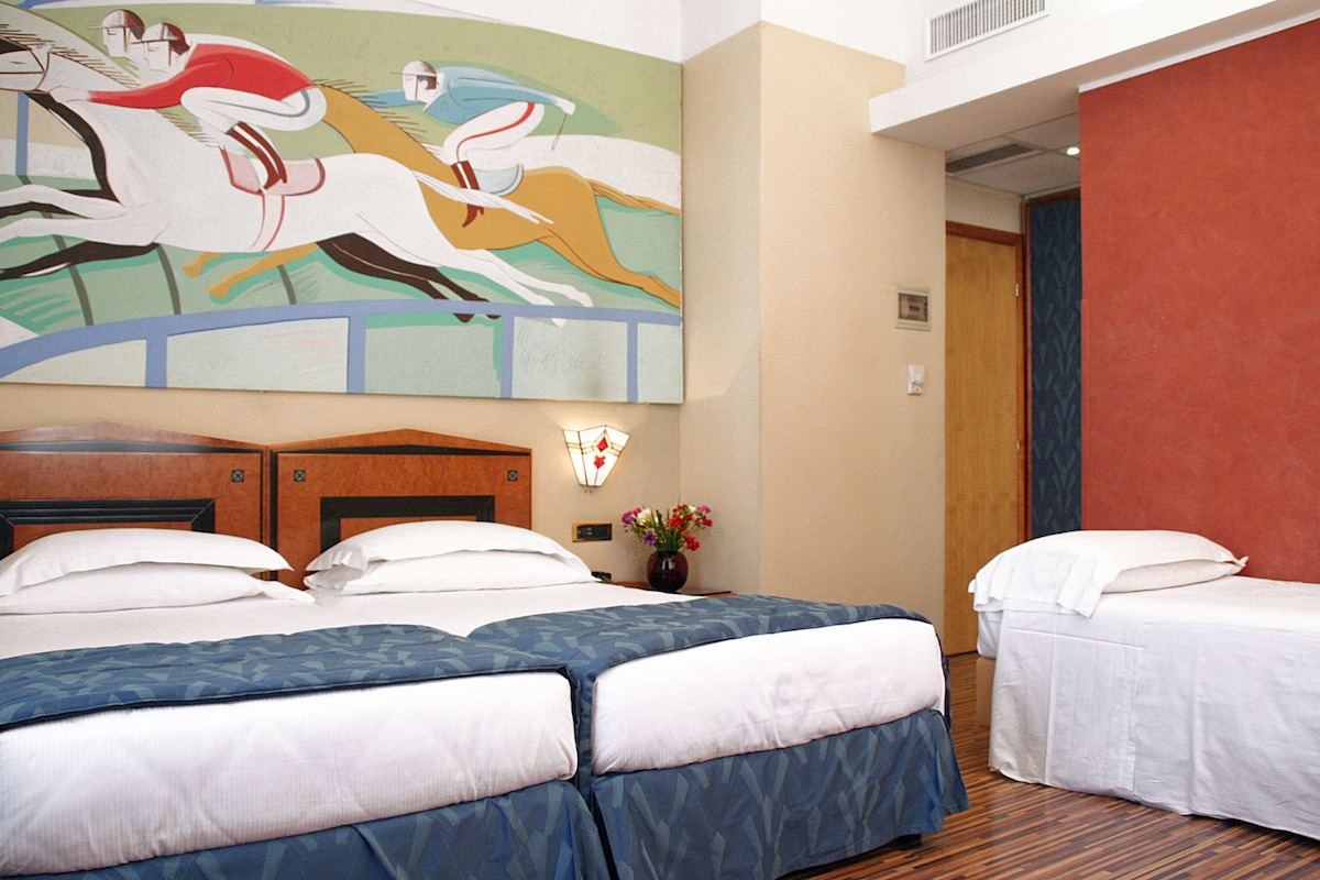 A hotel room with two beds, a large painting of horses, a vase with flowers, and a wooden headboard.