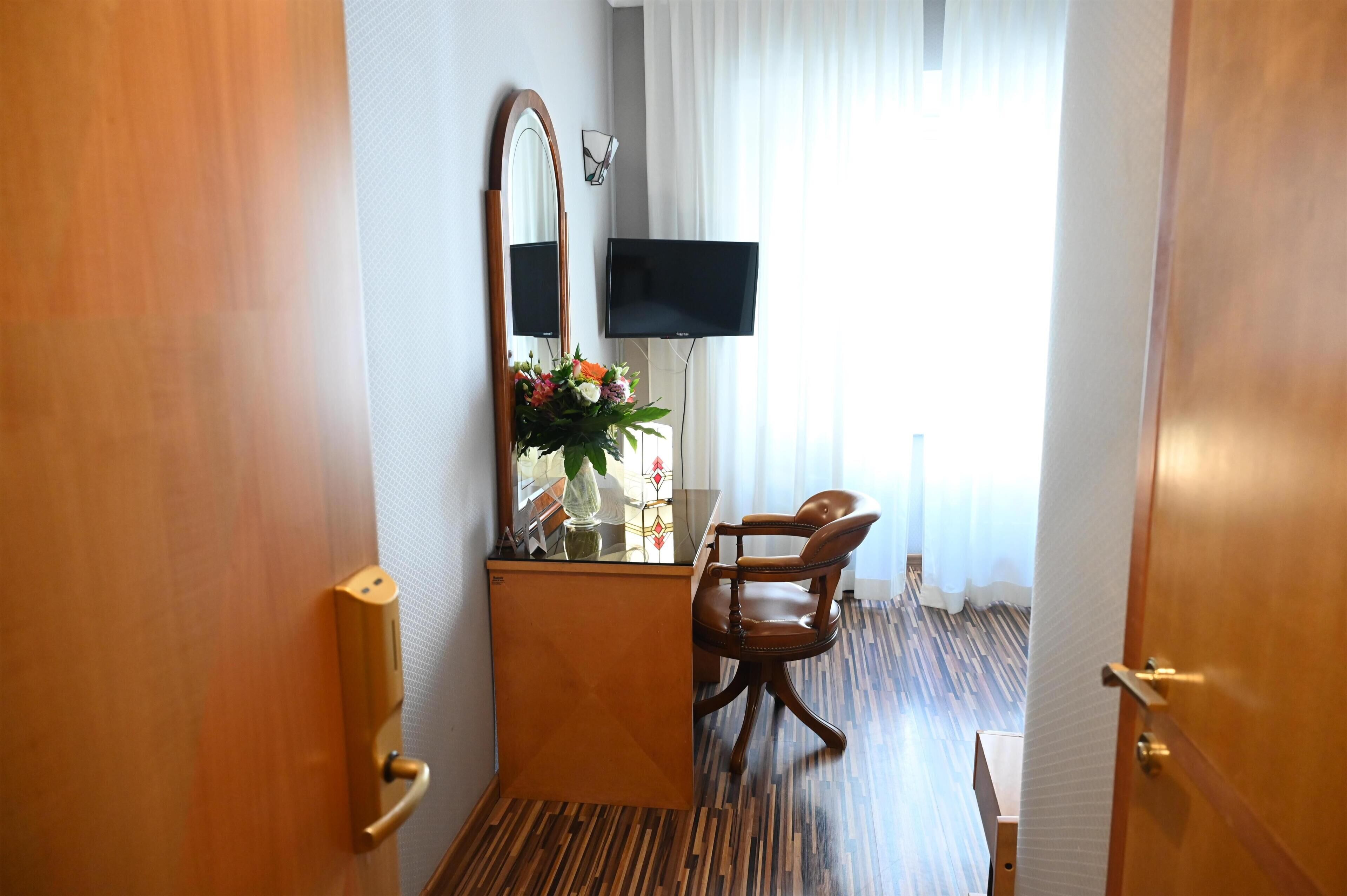 A hotel room with a wooden desk, a chair, a large mirror, a flat-screen TV, and a window with curtains.