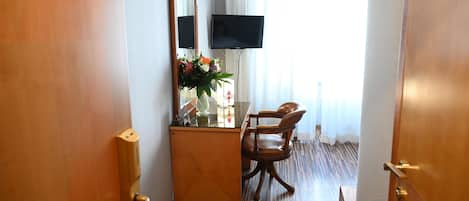 A hotel room with a wooden desk, a chair, a large mirror, a flat-screen TV, and a window with curtains.