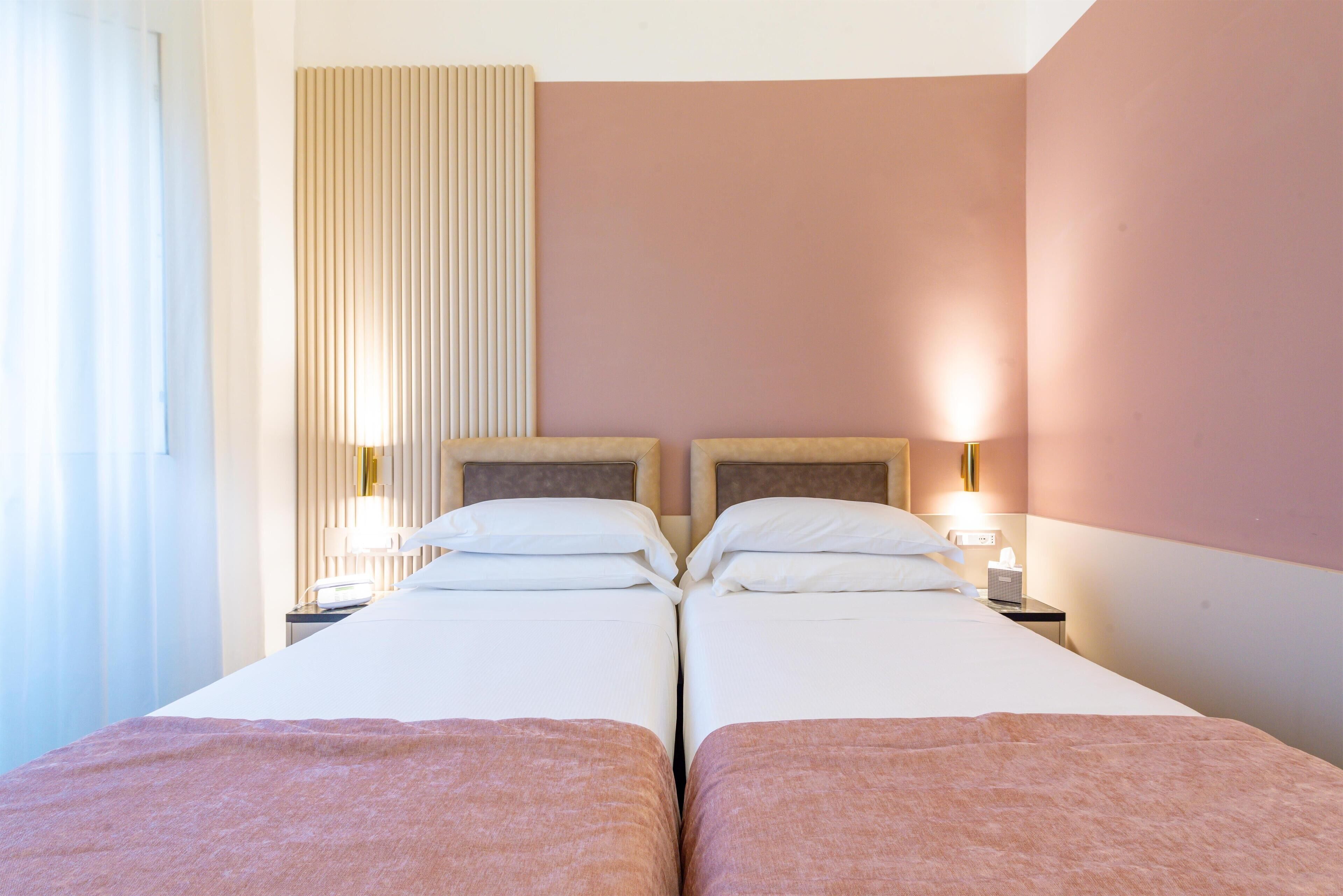 A hotel room with two beds, a bedside table with a lamp, and a window with curtains.