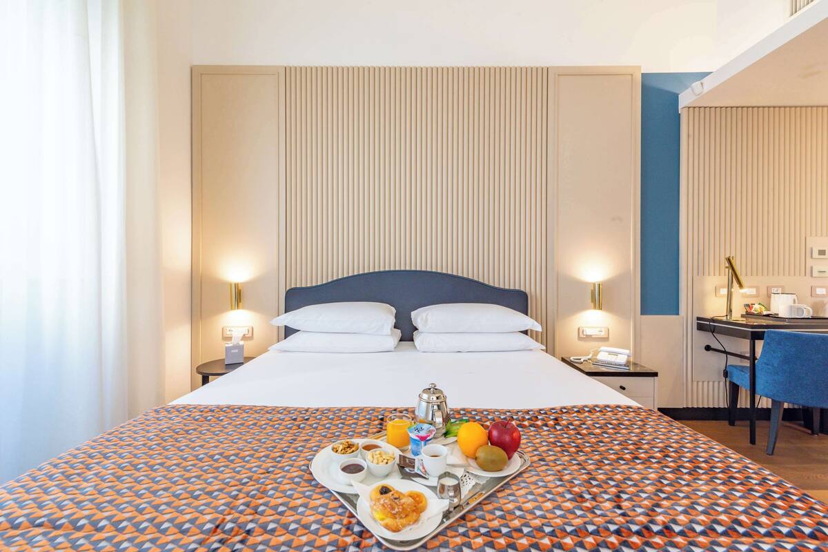 A hotel room with a bed, bedside tables, a desk, and a tray of breakfast items.