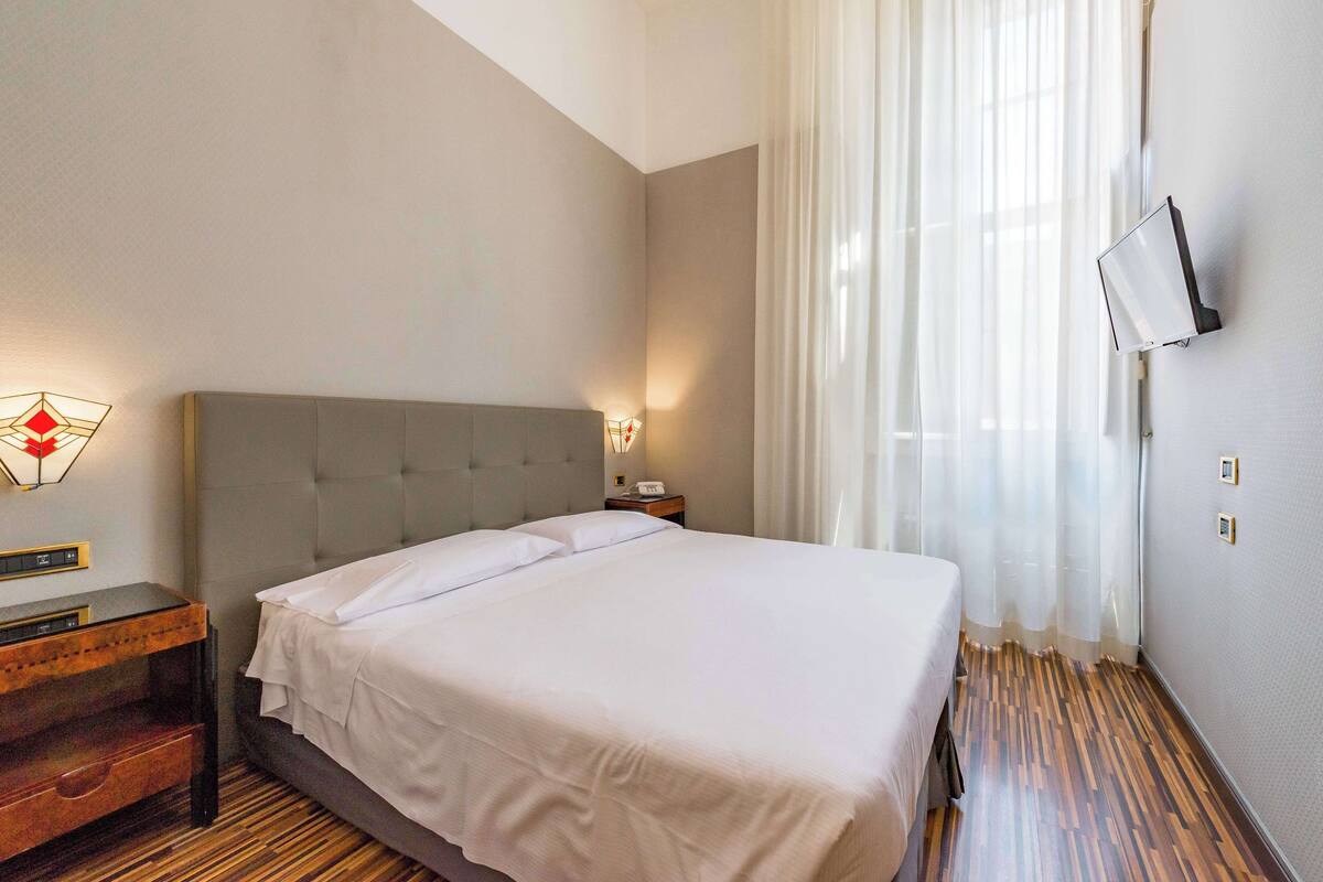Un lit bien fait dans une chambre d’hôtel, avec un plancher en bois, une table de chevet avec une lampe et un téléviseur fixé au mur.