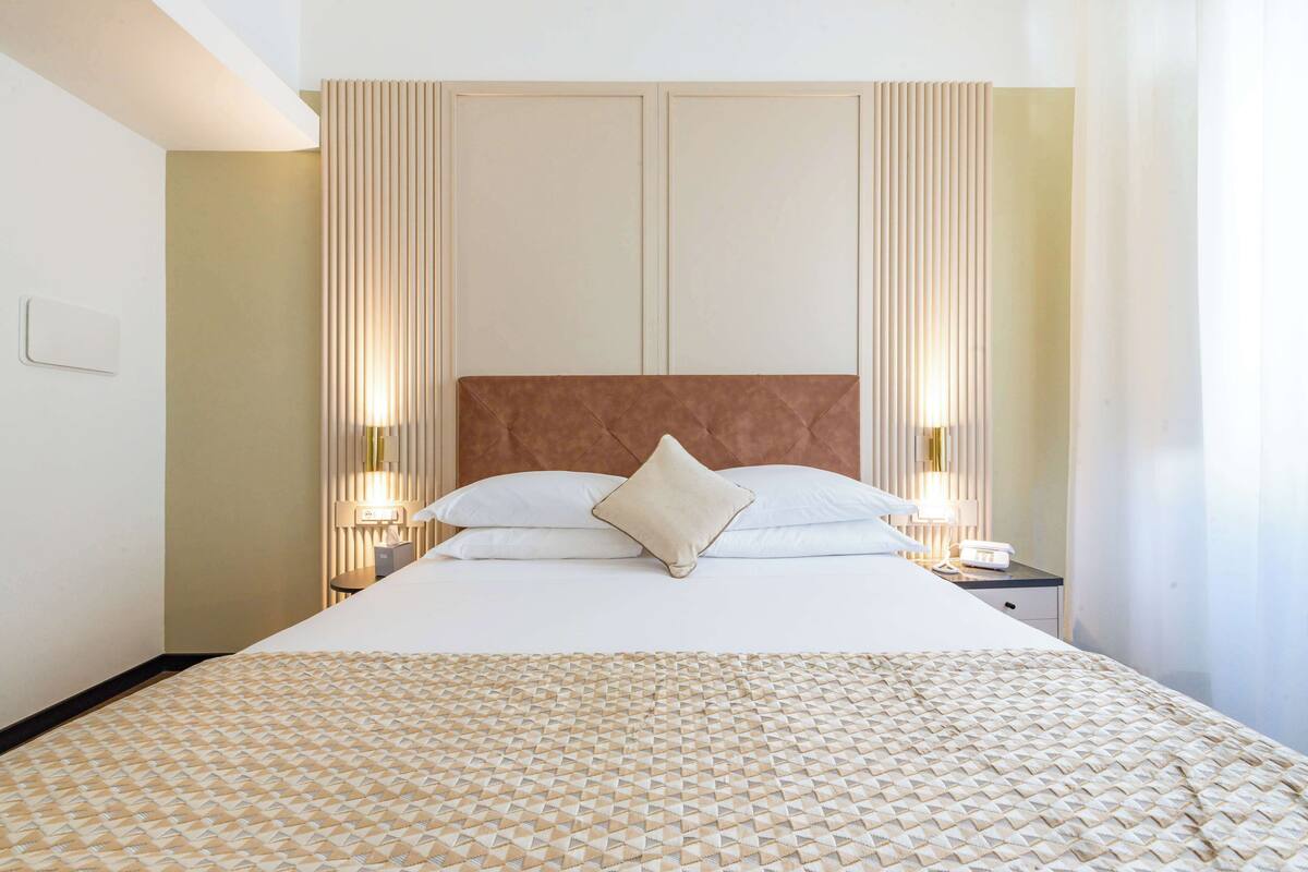 A hotel room with a large bed, two bedside tables with lamps, a headboard, and a wall-mounted light fixture.