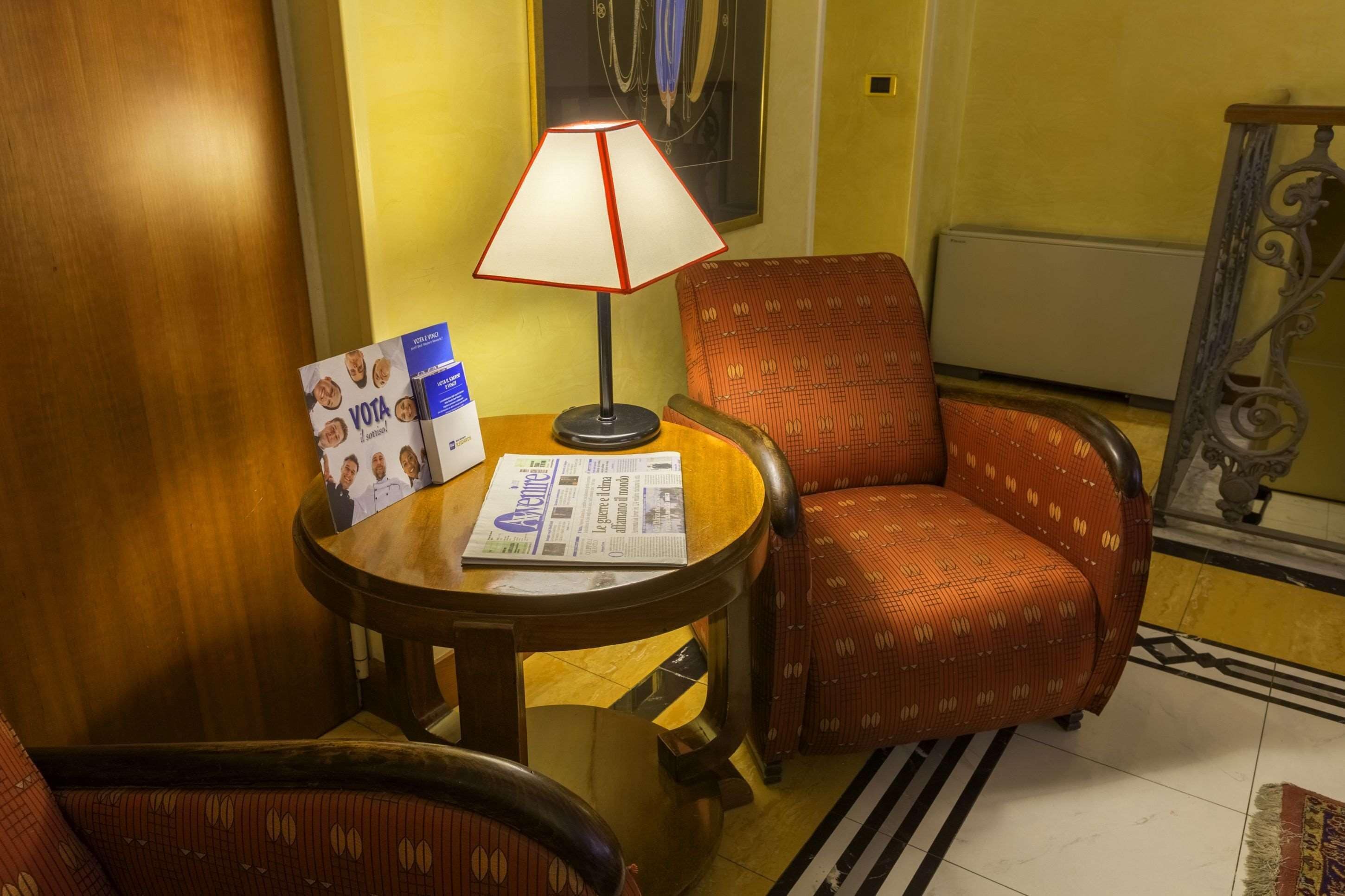 Ein Hotelzimmer mit einem Holztischchen, einem rot gemusterten Sessel, einer Lampe, einer Zeitung und einem gerahmten Bild an der Wand.