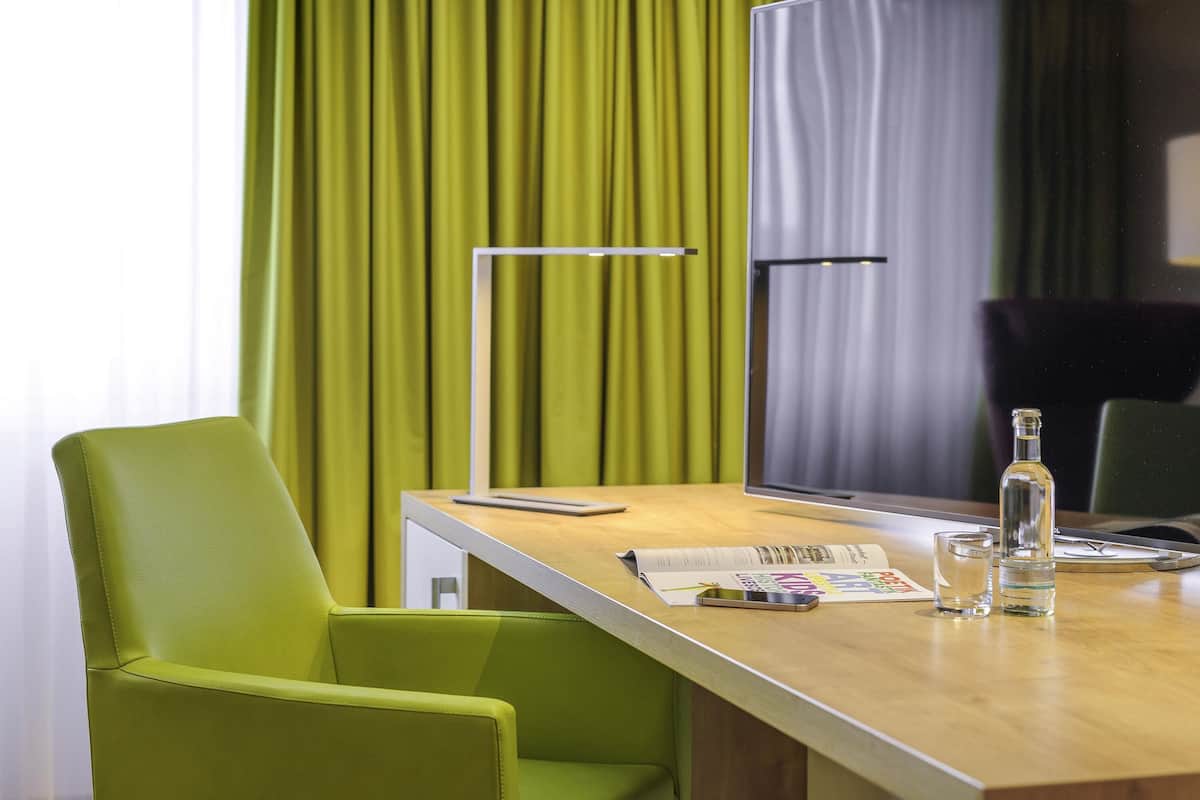 Ett hotellrum med ett skrivbord i trä, en grön stol, en platt-TV, en flaska och en tidning.