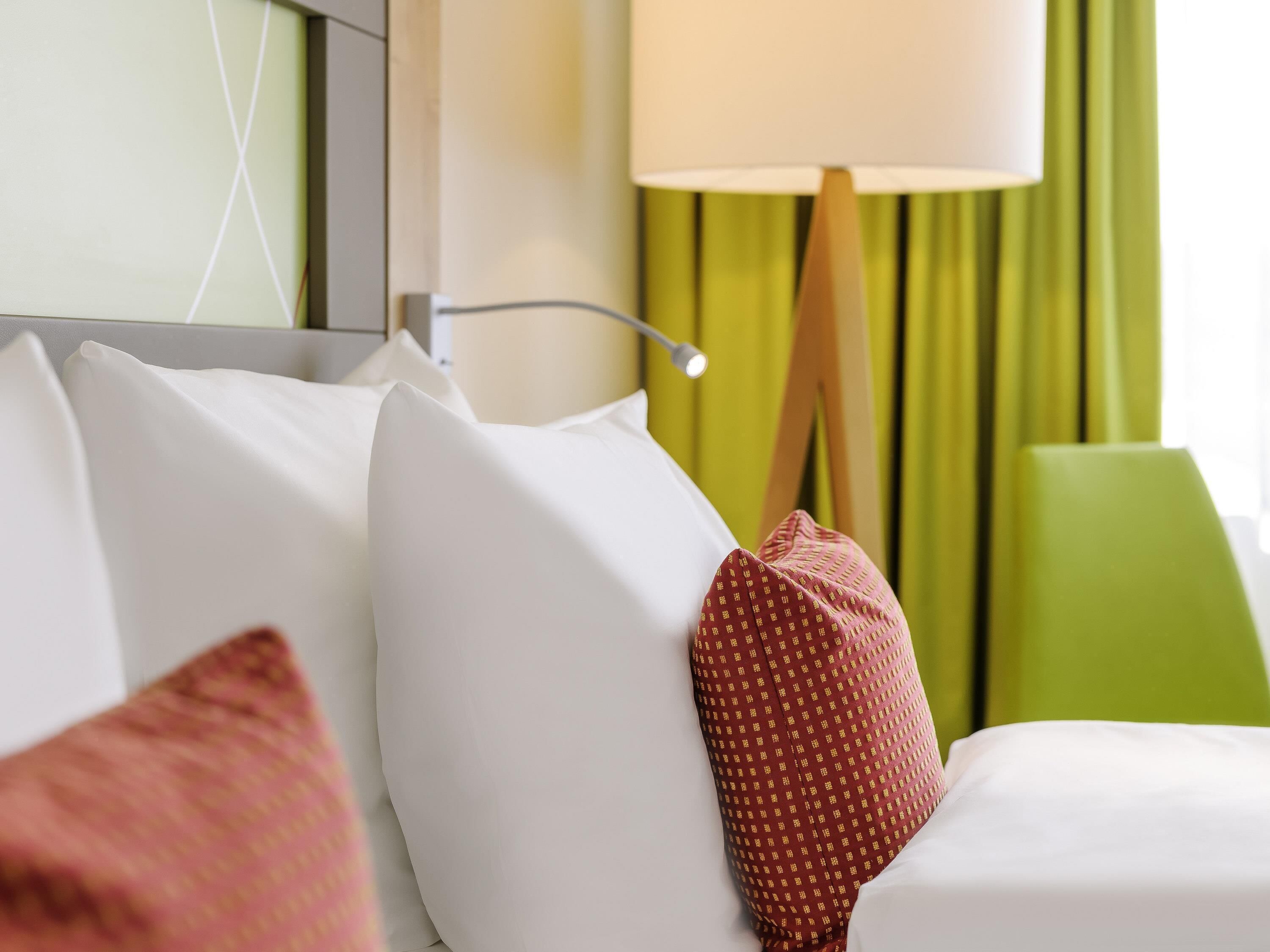 A hotel room with a bed, pillows, a lamp, and green curtains.