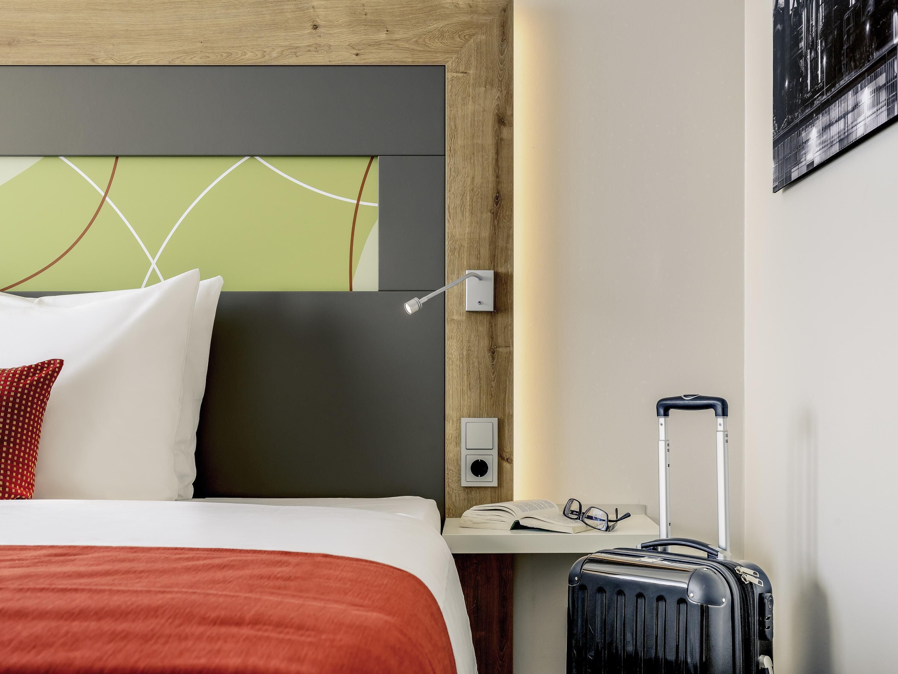 A hotel room with a bed, a nightstand, a suitcase, and a wall-mounted light.