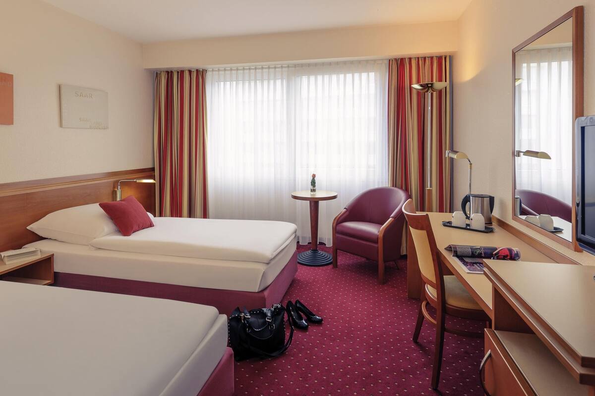 Une chambre d’hôtel avec deux lits, un bureau avec une chaise, une petite table avec une lampe, un miroir et une fenêtre avec des rideaux.