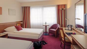 Une chambre d’hôtel avec deux lits, un bureau avec une chaise, une petite table avec une lampe, un miroir et une fenêtre avec des rideaux.