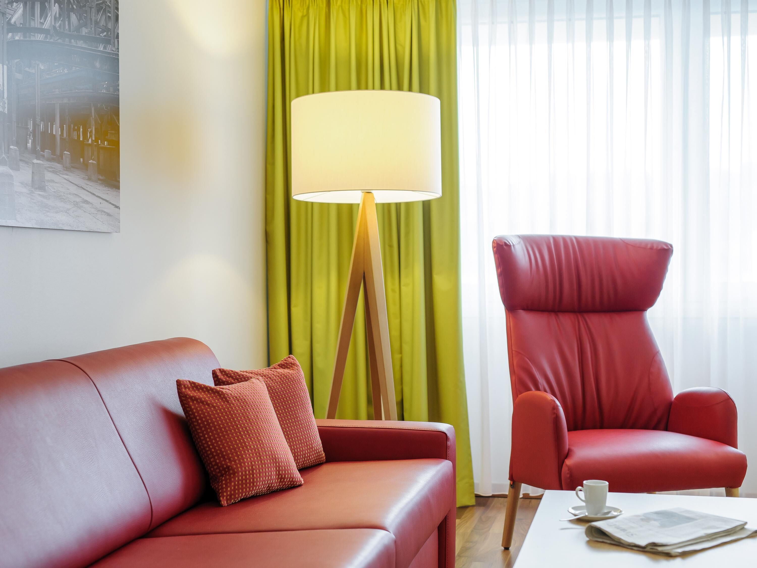 A red leather sofa with patterned cushions, a red armchair, a floor lamp, and a table with a cup and newspaper.
