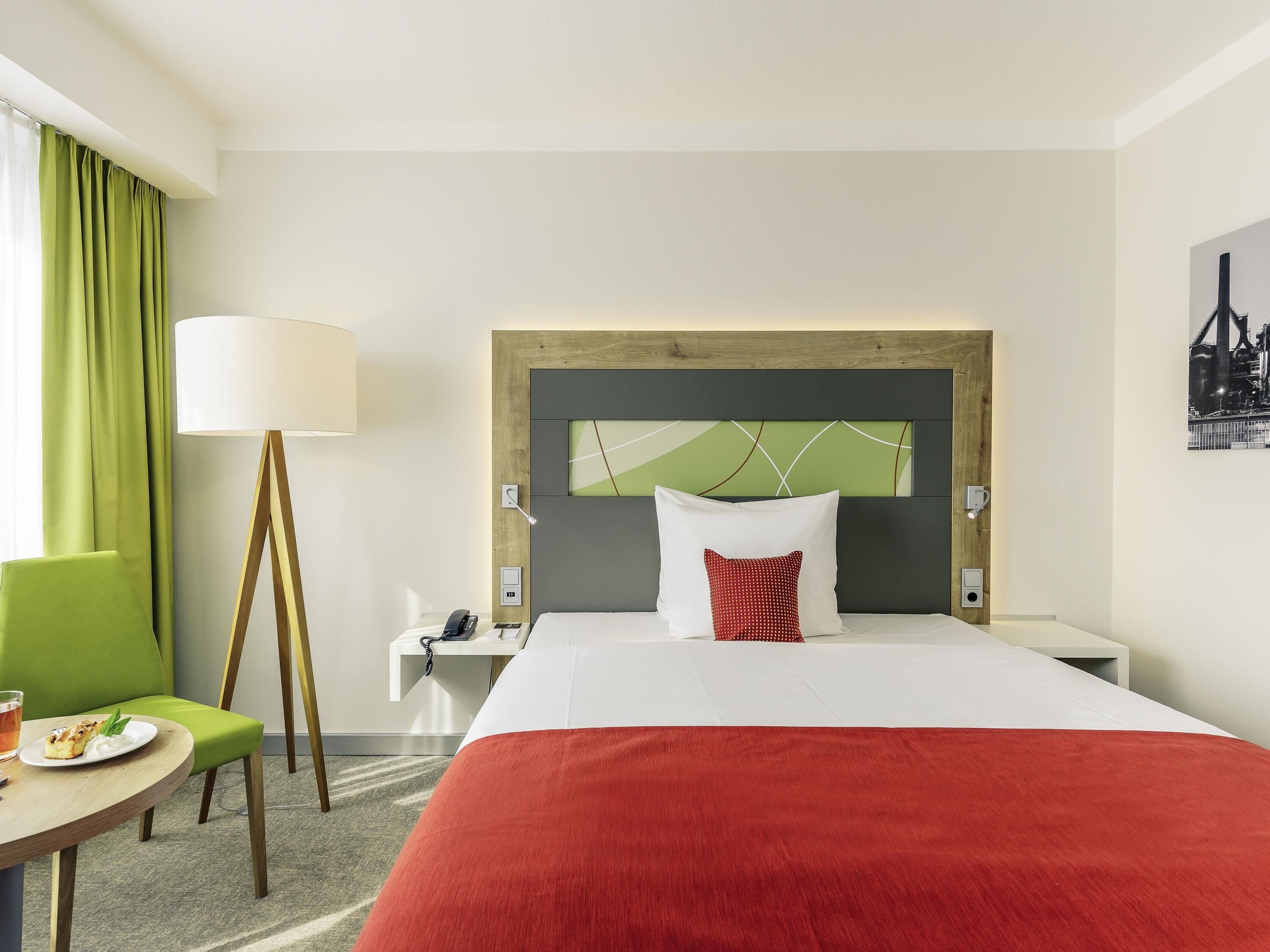 A hotel room with a bed, a desk, a chair, a lamp, and a painting on the wall.