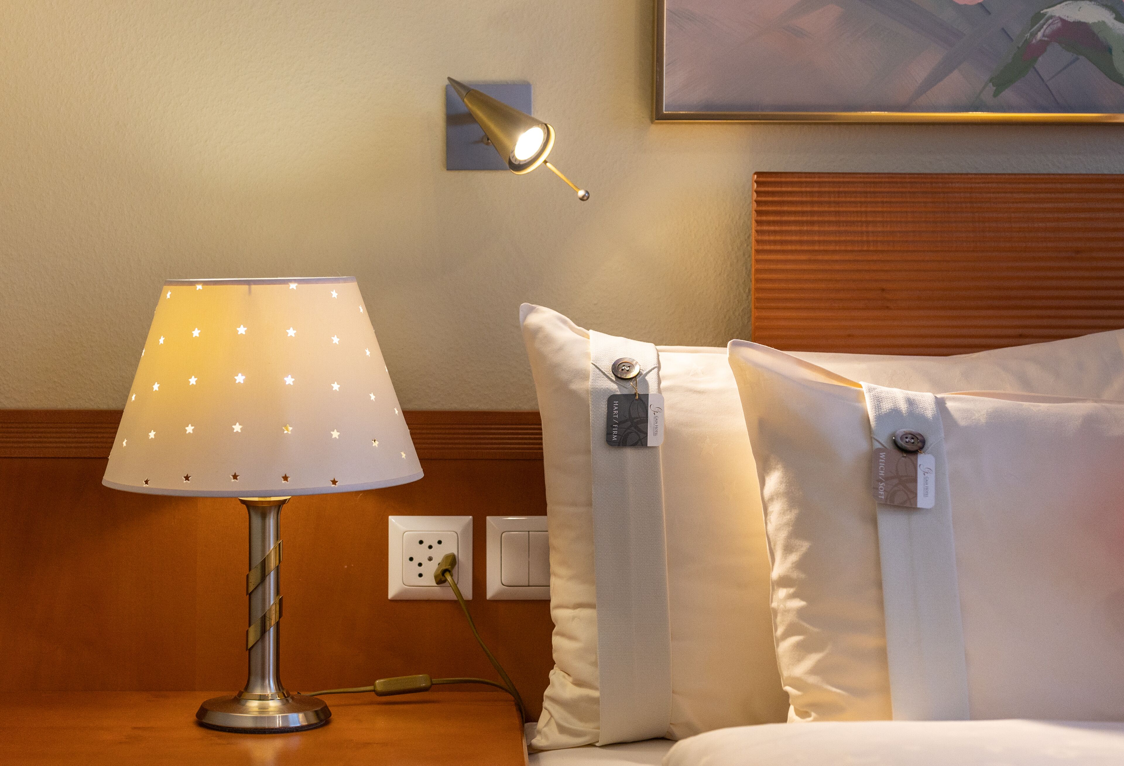 A hotel room with a bedside lamp, a nightstand, a power outlet, and a bed with pillows.