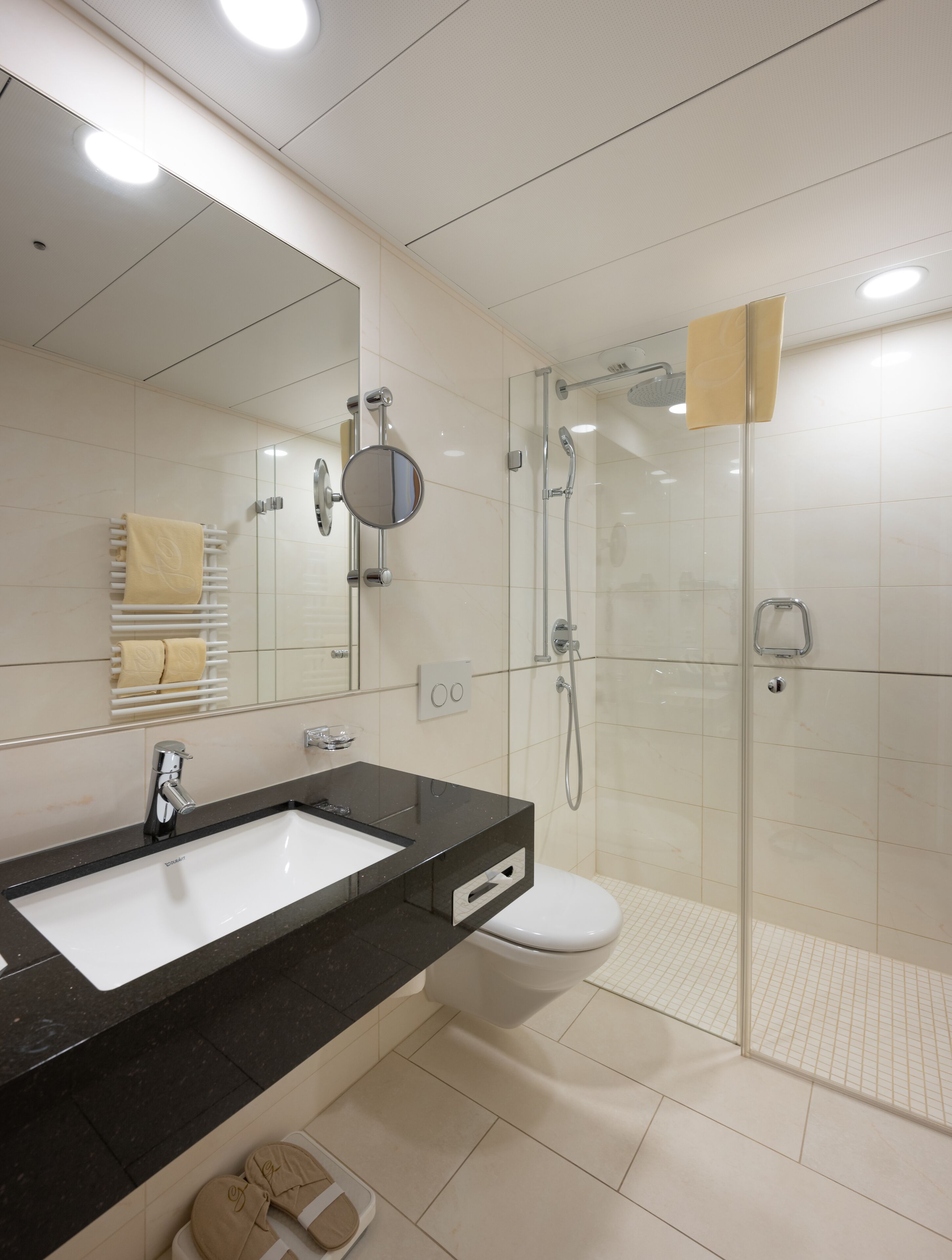 Une salle de bain moderne avec un lavabo sur un plan de toilette noir, des toilettes, une douche à parois de verre et un miroir surmonté d’un autre miroir rond.