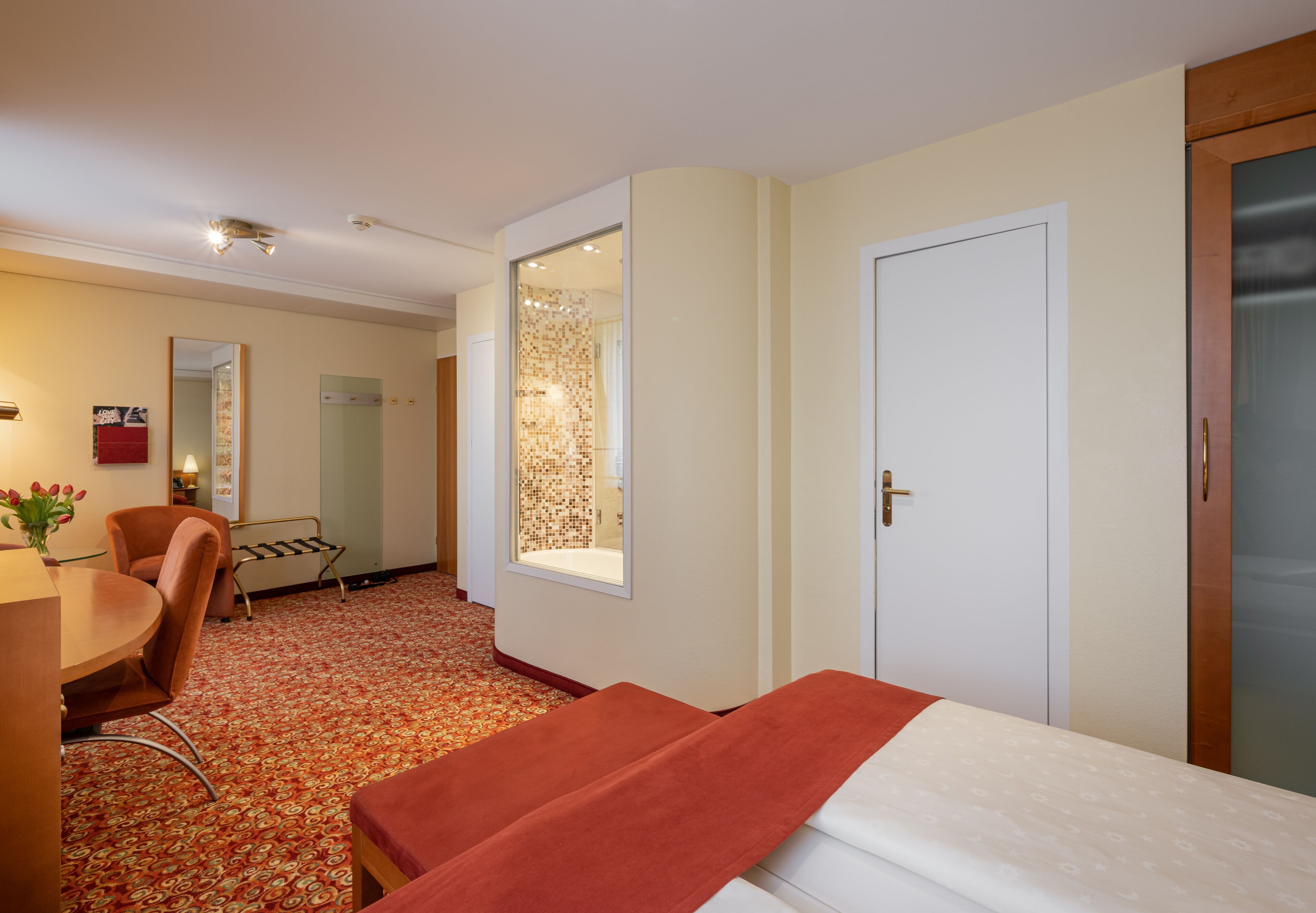 Habitación de hotel con cama, escritorio, silla, una pequeña mesa con flores, espejo y puerta.