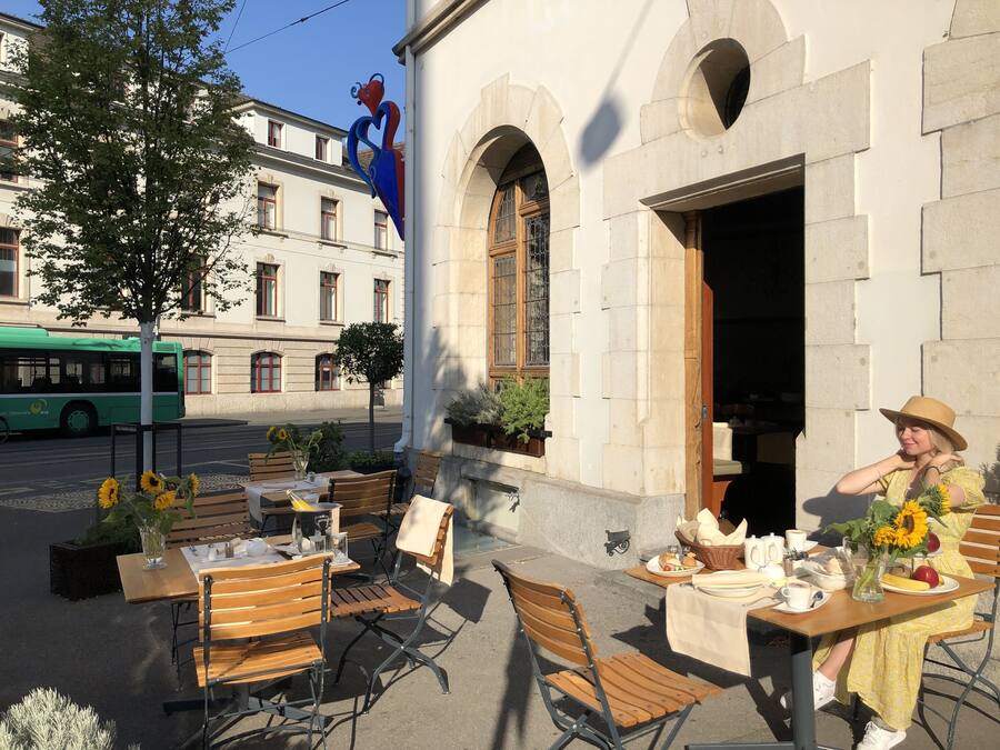 An outdoor café with tables and chairs, a green bus in the background, and a building with arched windows and a heart-shaped sculpture.