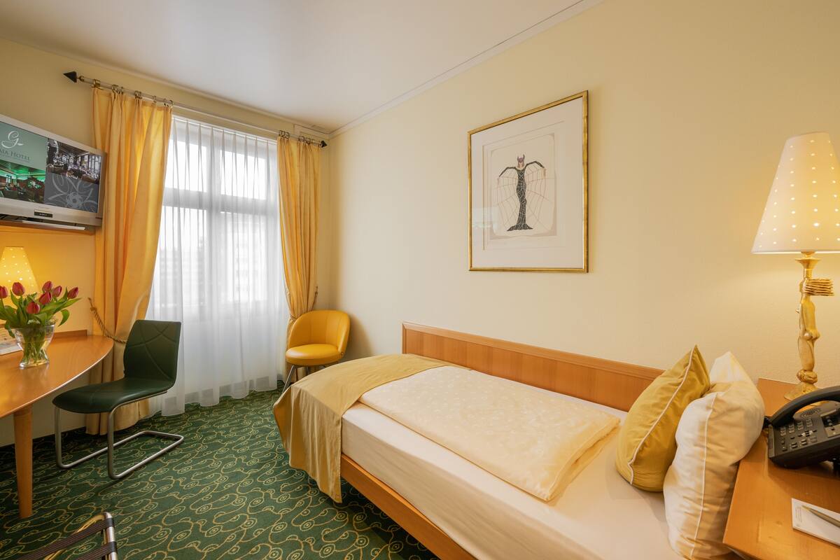A hotel room with a bed, a desk with a chair, a TV, a framed picture on the wall, and a lamp.
