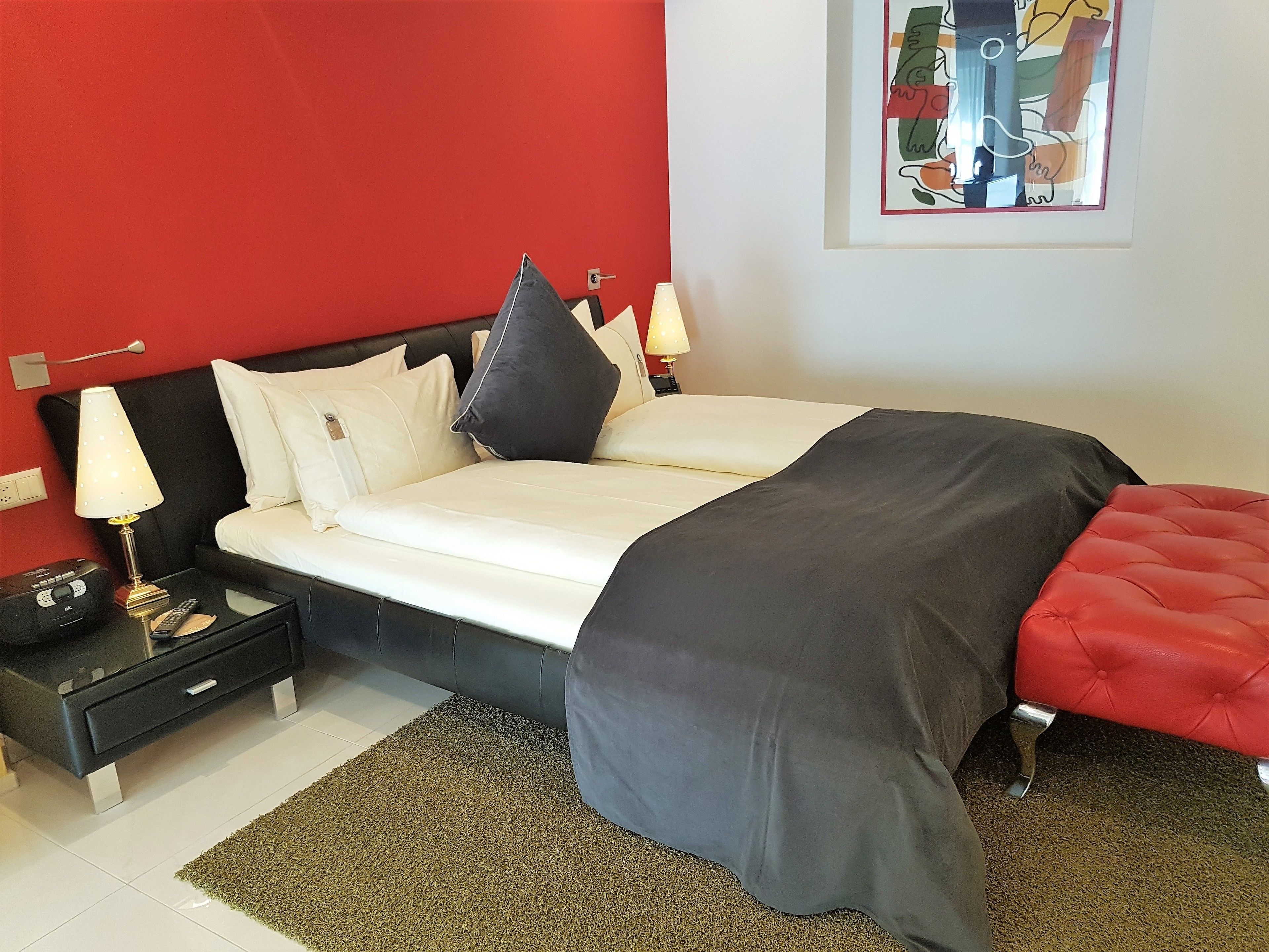 Ein modernes Schlafzimmer mit roter Akzentwand, einem großen Bett mit schwarz-weißem Überwurf, einem roten Hocker, einem Nachttisch mit Lampe und einem Bilderrahmen mit einem abstrakten Gemälde.