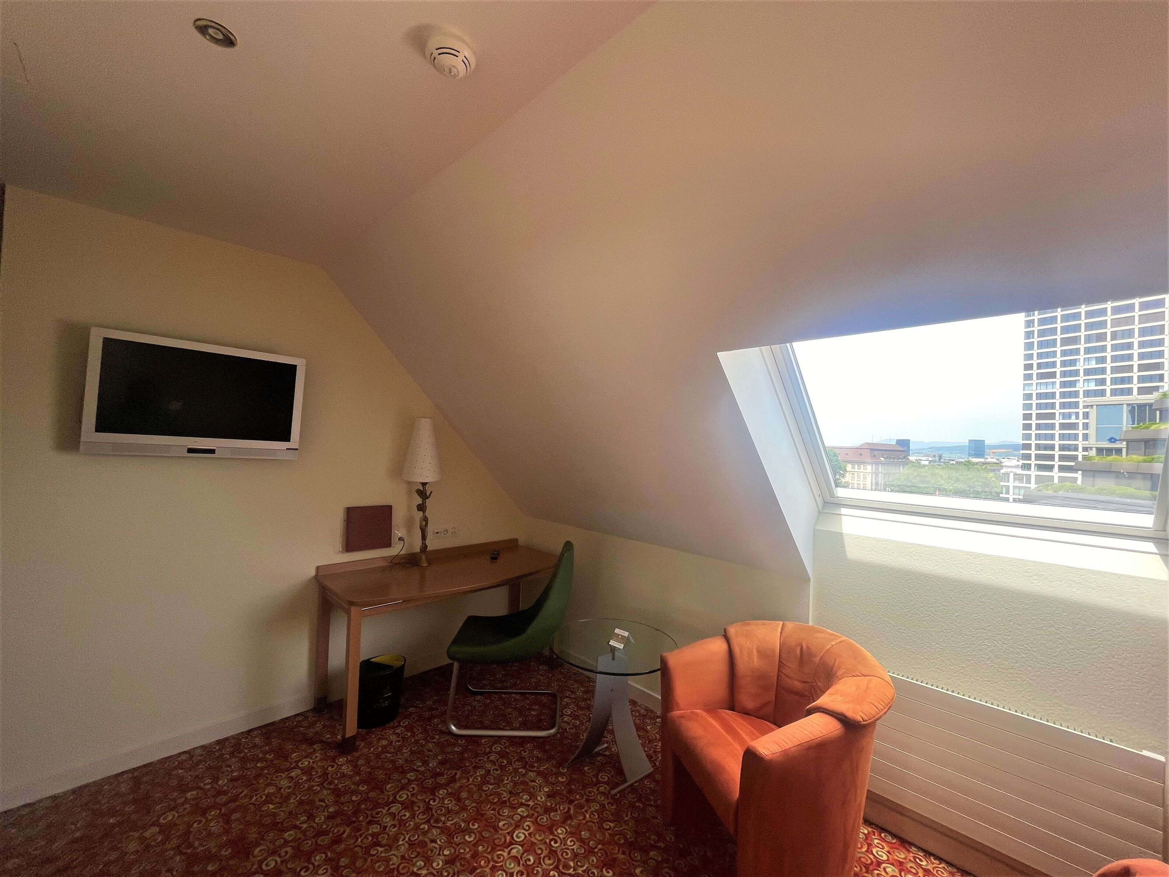 A small, triangular room with a television, a desk, a chair, and a large window offering a view of buildings.