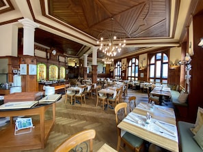 A spacious dining area with wooden tables and chairs, large windows, and a high ceiling with wooden beams.