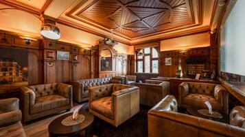 A spacious, elegantly furnished lounge area with leather sofas and armchairs, wooden paneling, and a high ceiling with a geometric design.