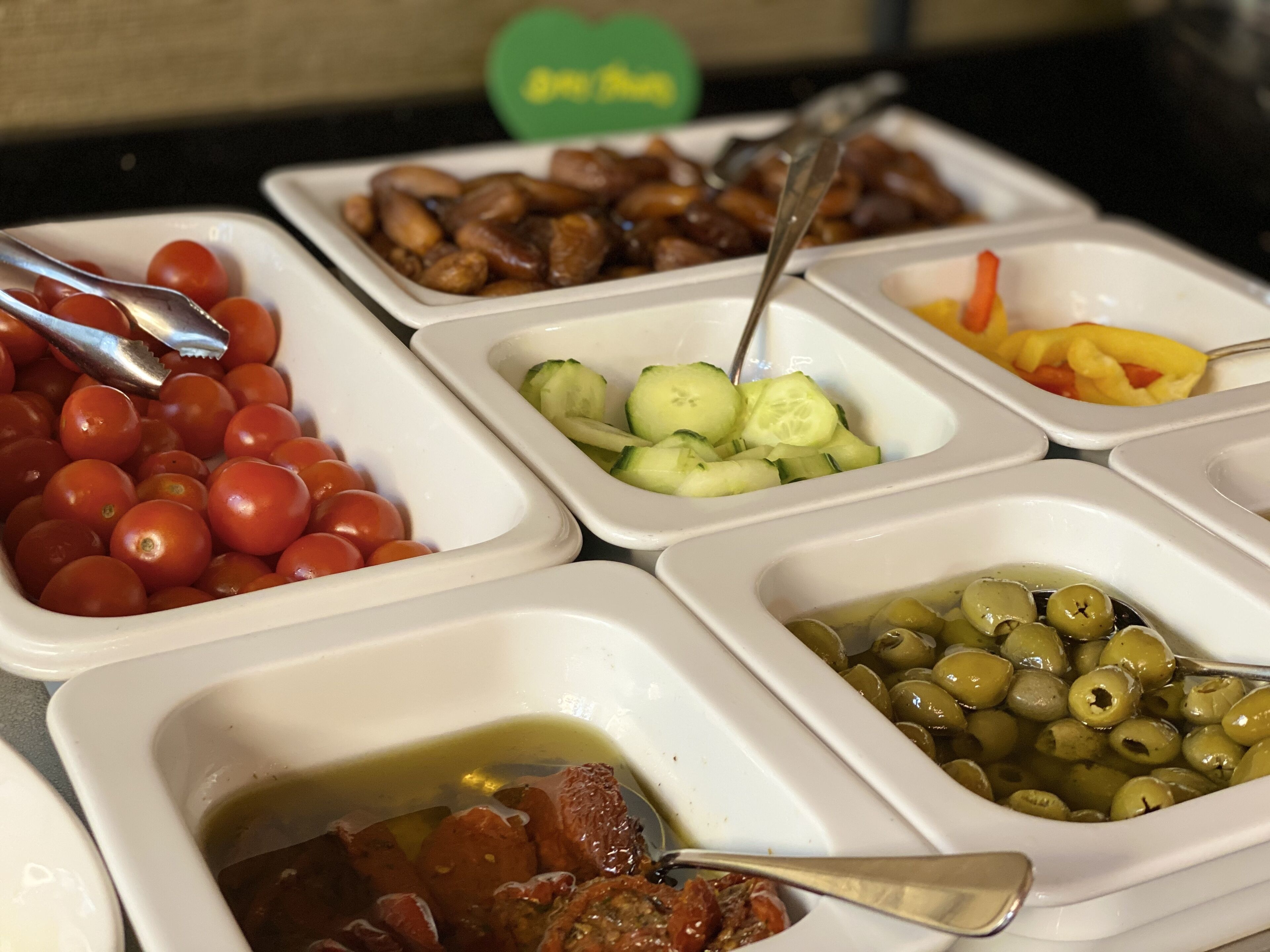 Een buffet met diverse gerechten, waaronder tomaten, komkommers, olijven, en een schotel met groene saus.