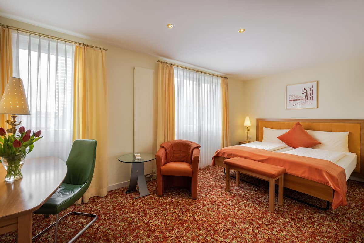 A hotel room with a bed, a chair, a small table, and a desk with a lamp and flowers.