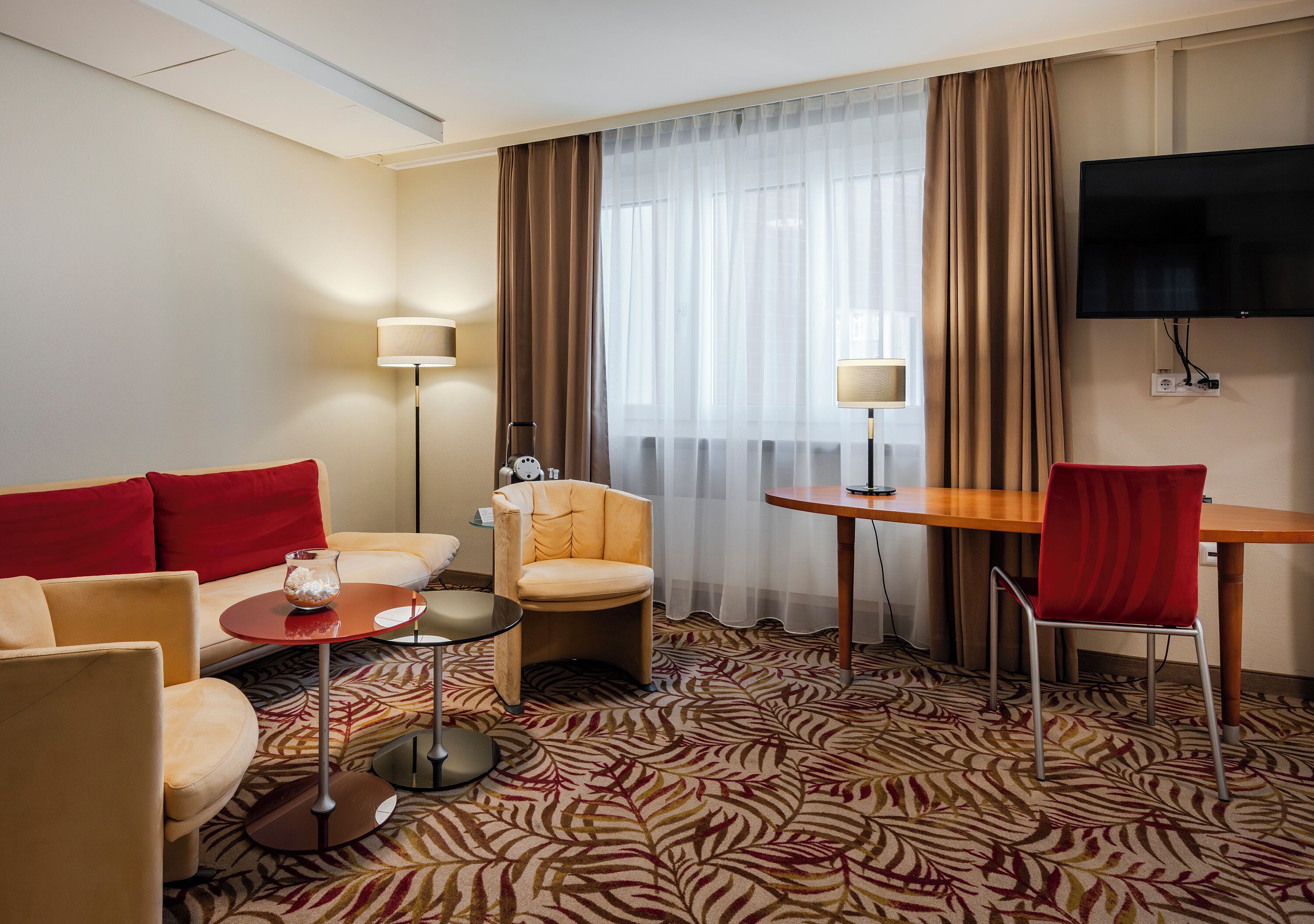 A hotel room with a sofa, armchairs, a small table, a TV, and a patterned carpet.