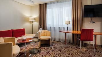 A hotel room with a sofa, armchairs, a small table, a TV, and a patterned carpet.