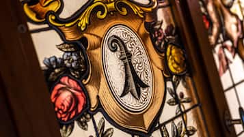 A stained glass window with a shield-shaped design, featuring a prominent letter 'R' and floral patterns.