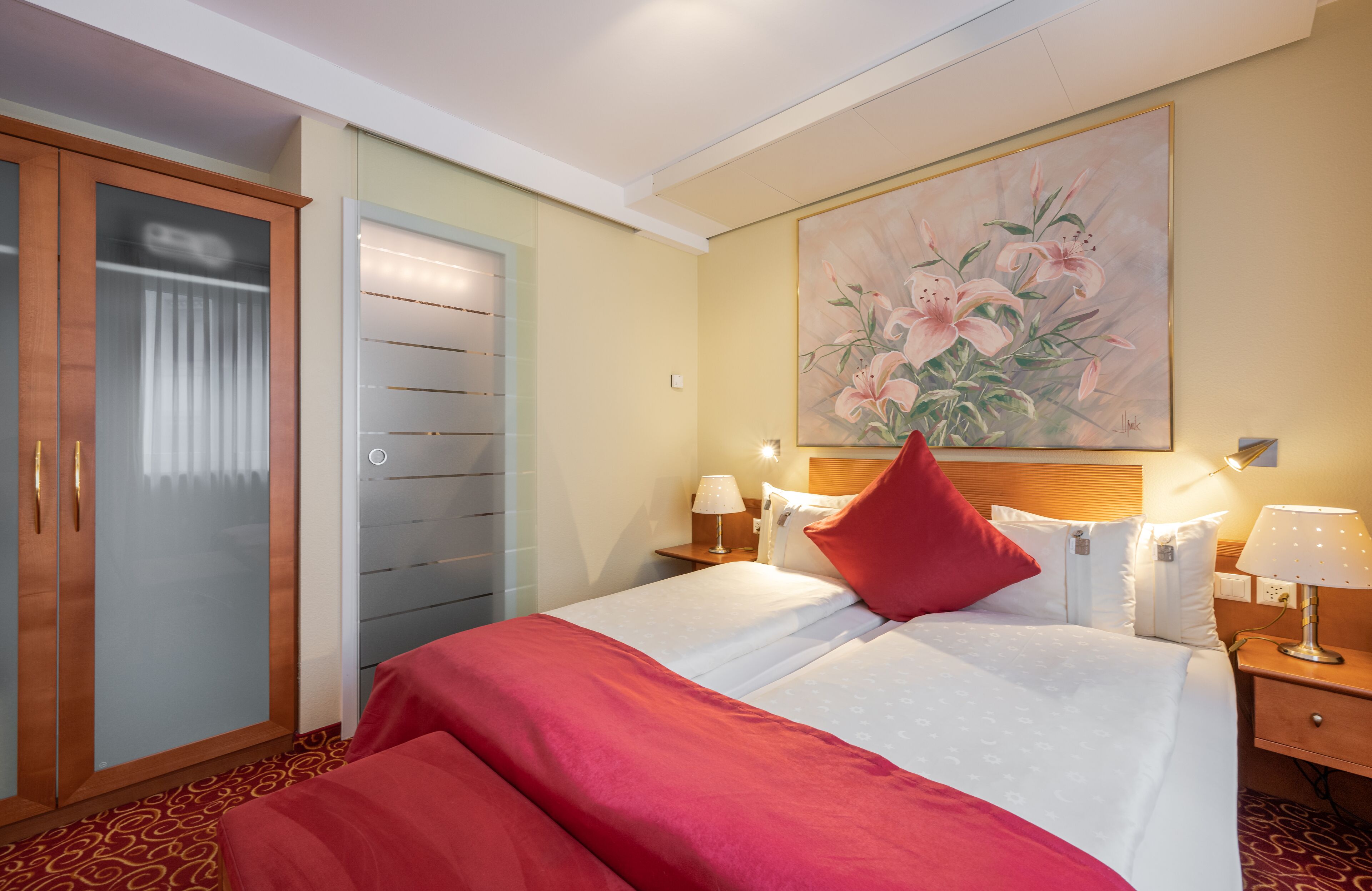 A hotel room with a bed, a wardrobe, a painting, and two bedside lamps.