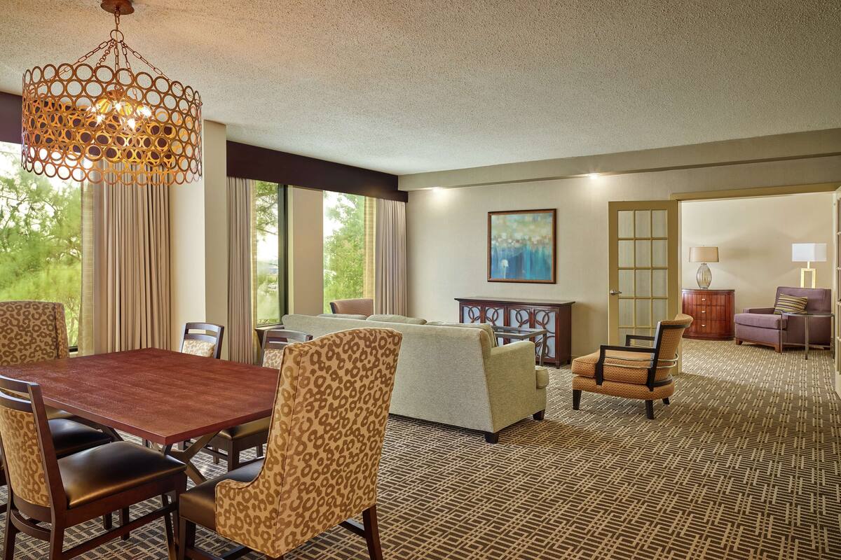 Ein Hotelzimmer mit Essbereich, einer Sofagarnitur, einem gemusterten Teppich und einem großen Fenster mit Vorhängen.