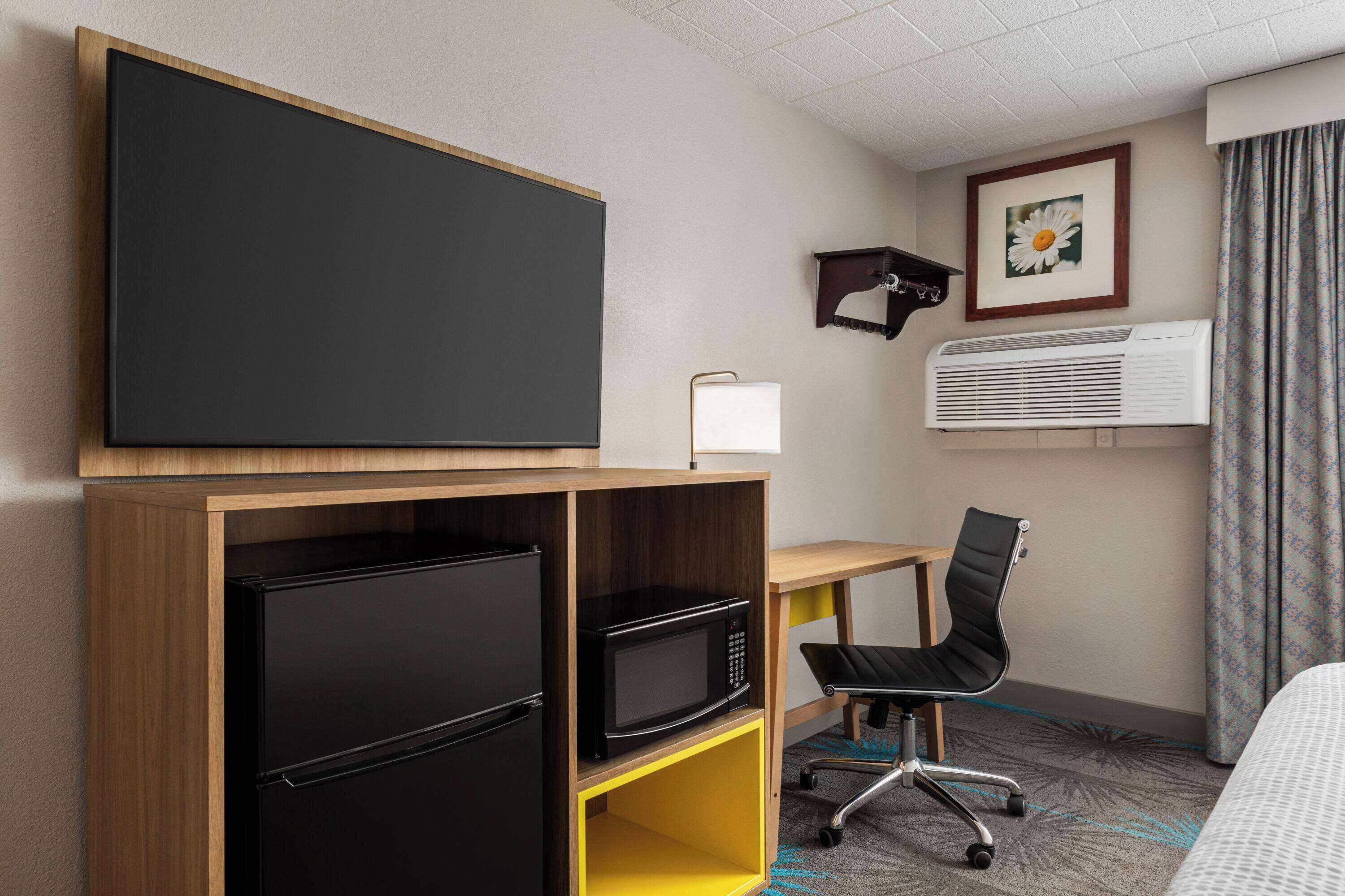 A hotel room with a bed, a desk with a chair, a black refrigerator, a microwave, a flat-screen TV, an air conditioner, and a framed picture on the wall.