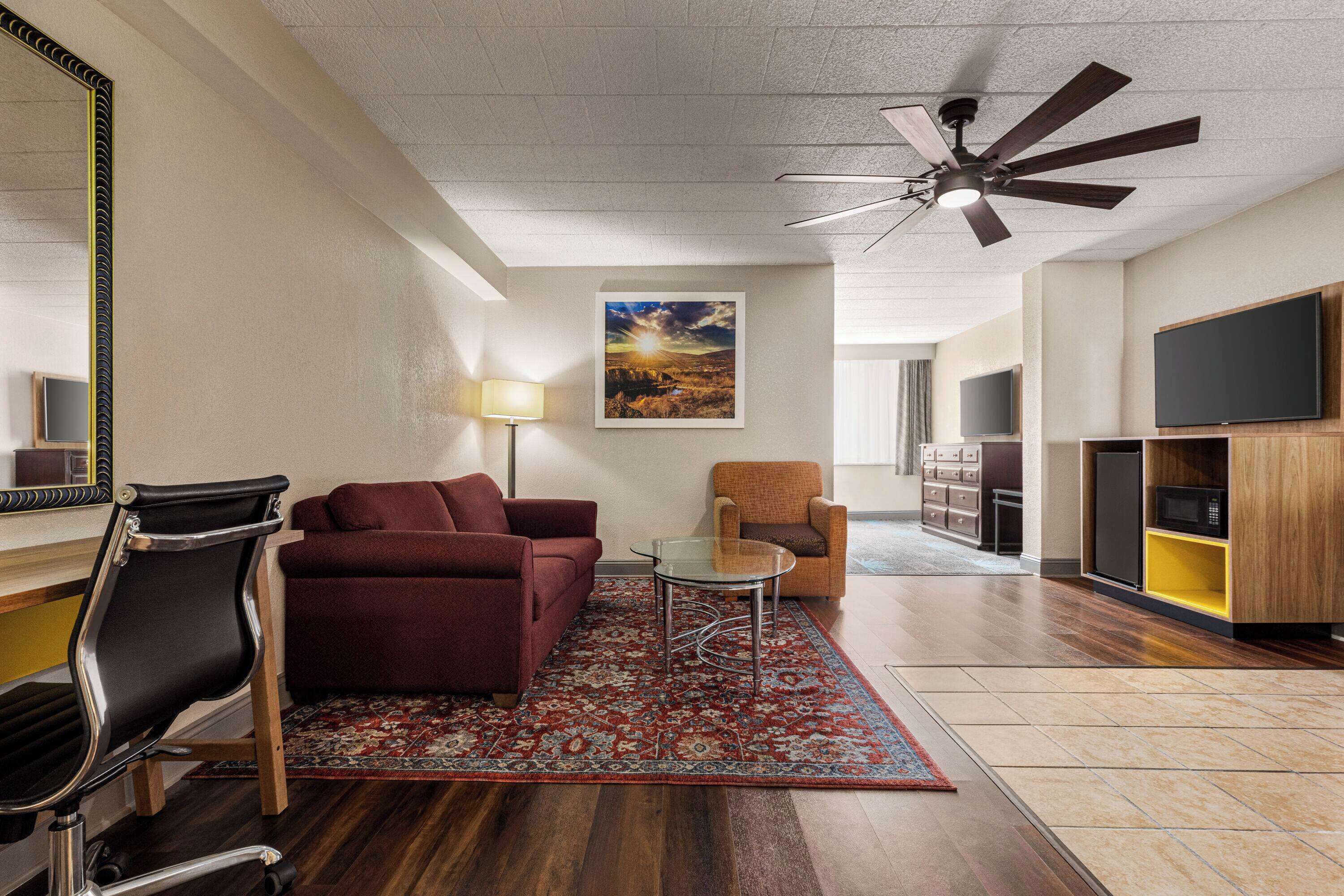A modern living room with a sofa, armchair, and a glass coffee table. There is a ceiling fan, a flat-screen TV, and a wooden cabinet.