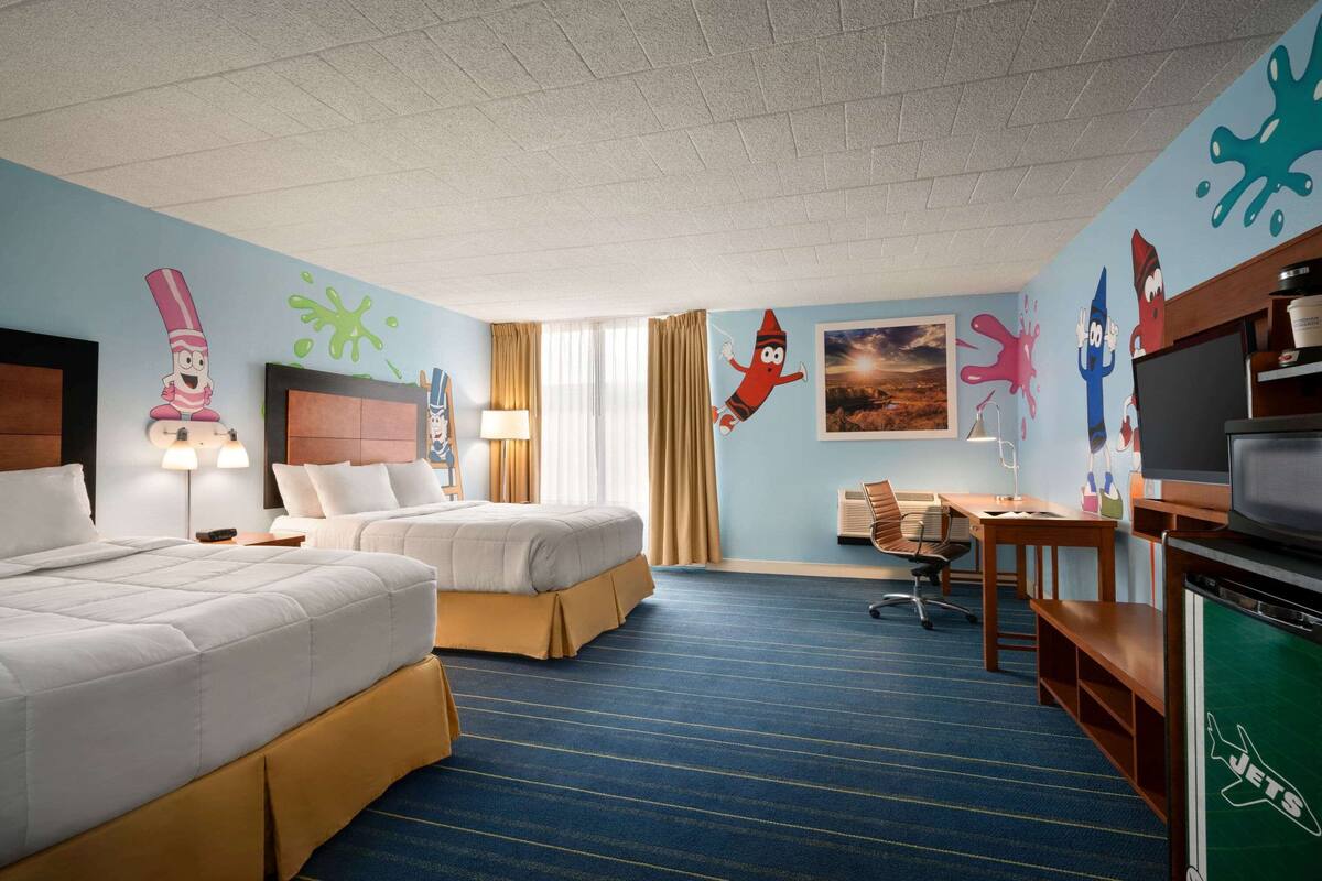 Ein Hotelzimmer mit zwei Betten, einem Schreibtisch, einem Fernseher und farbenfroher Wandgestaltung.