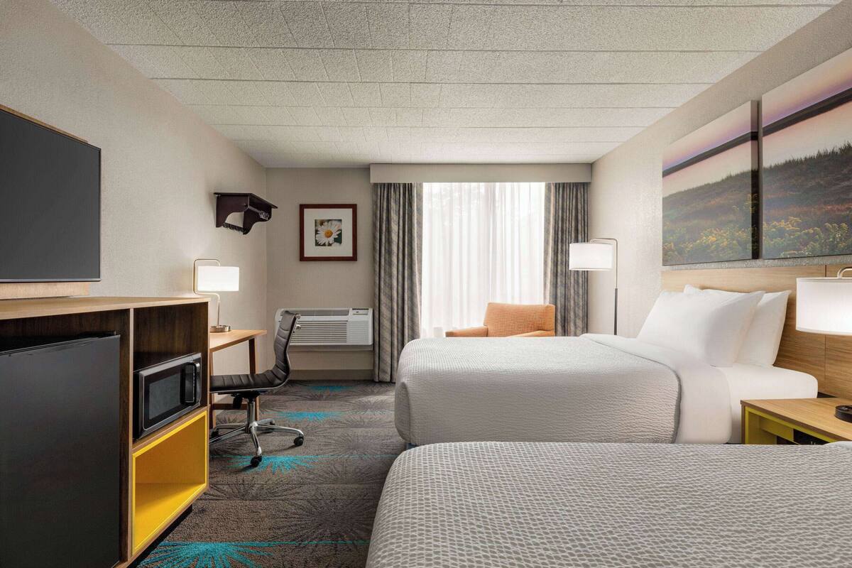 Ein Hotelzimmer mit zwei Betten, einem Schreibtisch mit Computer, einem Stuhl, einem Fernseher und einem Fenster mit Vorhängen.