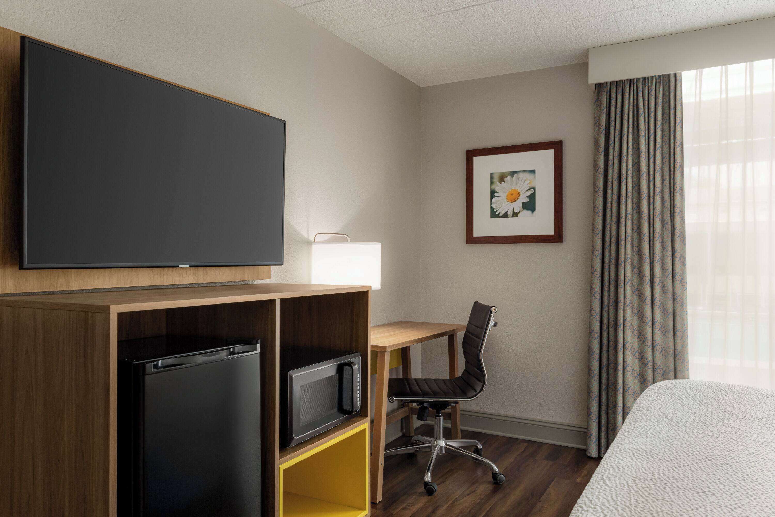 A hotel room with a bed, a desk with a chair, a television, a microwave, and a framed picture on the wall.
