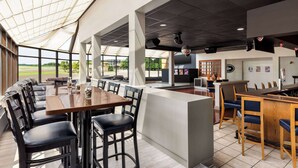 Un restaurante espacioso con techo alto, ventanas grandes y una variedad de zonas de estar, incluyendo una barra y una mesa con sillas.